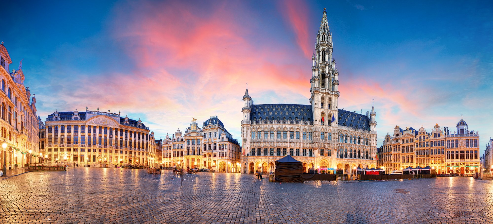 A stunning twilight view of Brussels' iconic Grand Place (Grote Markt) featuring the ornate Gothic Town Hall with its distinctive spire, surrounded by elegant 17th-century guild houses illuminated by warm golden lights against a vibrant blue and pink sunset sky. The cobblestone plaza is wet from rain, reflecting the architectural splendor of Belgium's most famous square.