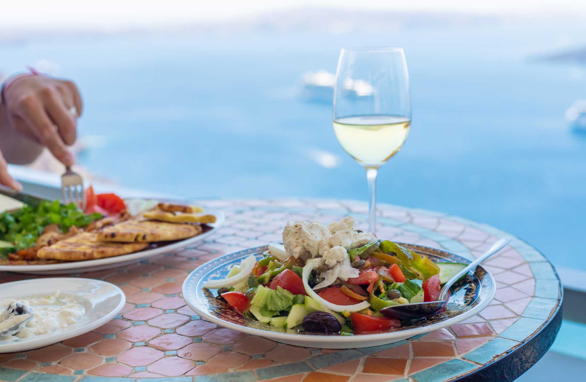 A scenic Mediterranean dining scene featuring a fresh Greek salad with feta cheese, olives, and vegetables on a decorative plate, paired with a glass of white wine. The table overlooks a stunning blue sea and sky, capturing the essence of Greek island hospitality and coastal leisure dining.
