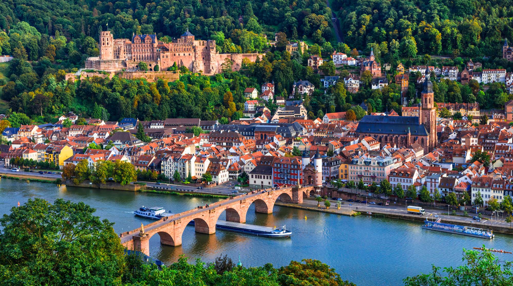 An aerial view of Heidelberg, Germany, showcasing the iconic red sandstone Karl Theodor Bridge spanning the Neckar River, with the medieval Heidelberg Castle perched on the forested hillside above the charming old town's terracotta-roofed buildings and historic church spires.
