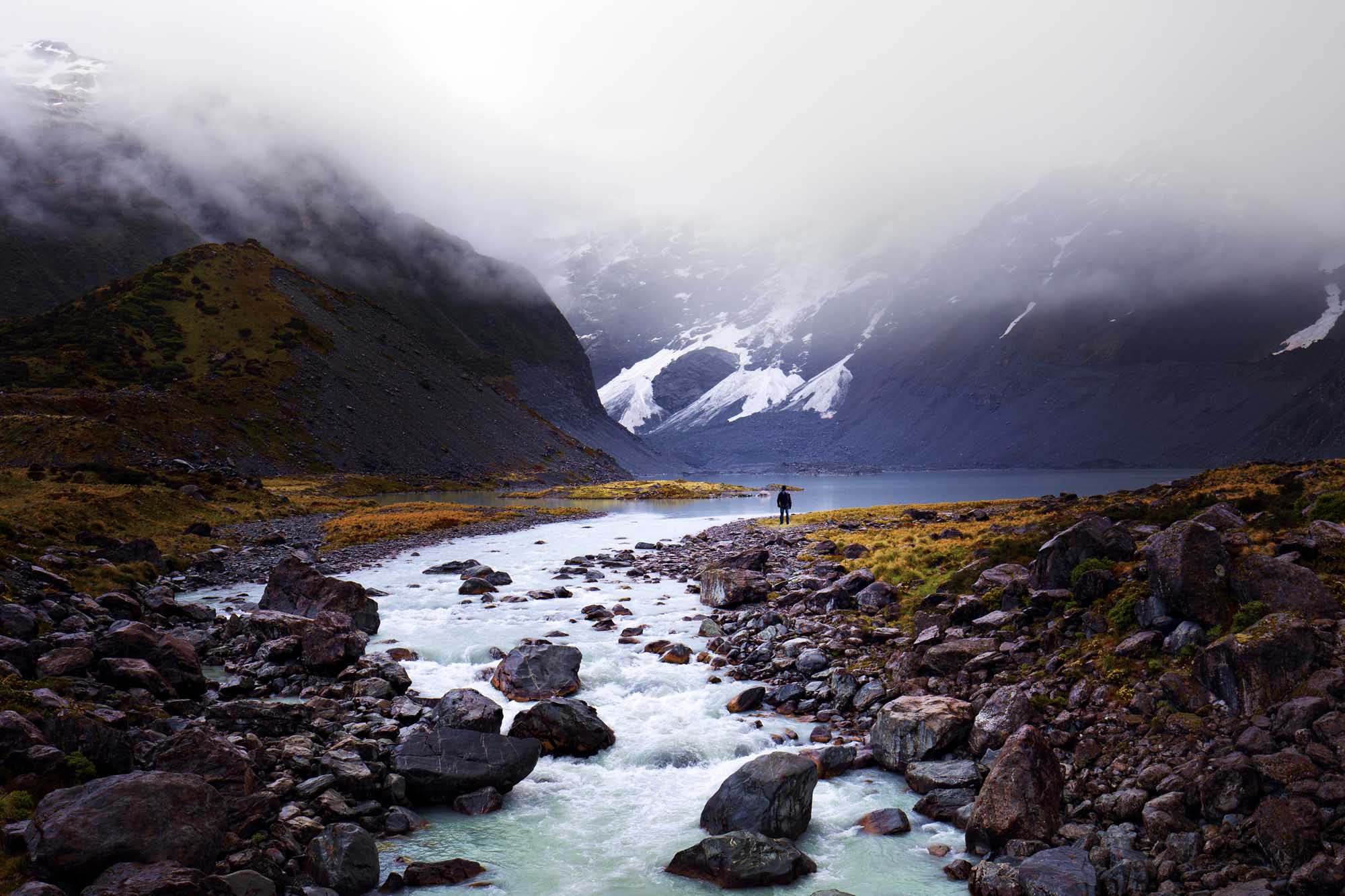 Hiker in Arctic Mountain Valley with Glacier A solitary hiker stands in a remote Arctic valley surrounded by snow-capped mountains shrouded in mist, with a pristine glacial stream flowing across rocky terrain in the foreground. The dramatic landscape features towering peaks with waterfalls and glaciers visible through the fog, creating a stark and otherworldly wilderness scene.