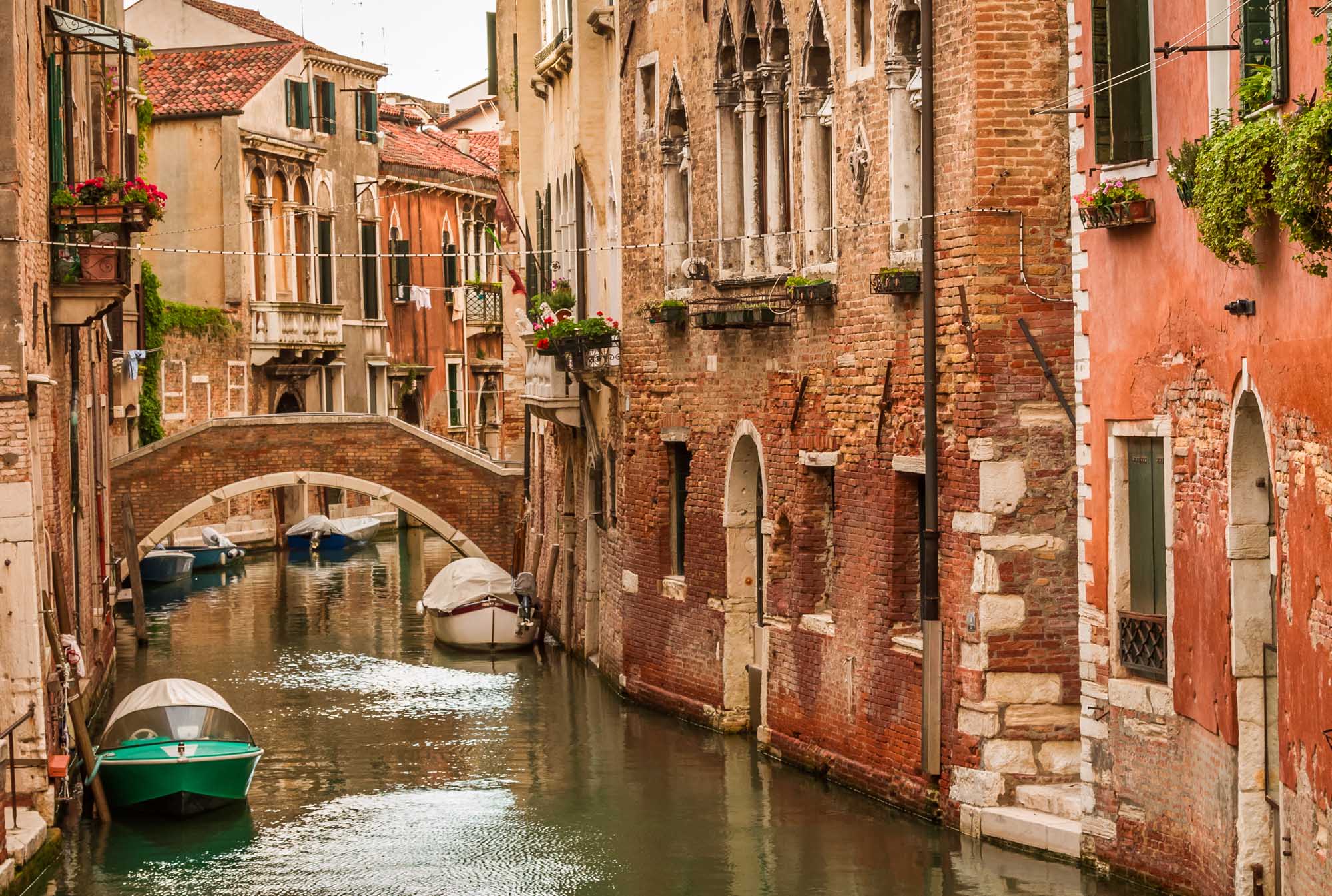 A picturesque view of a narrow Venetian canal lined with historic brick and stone buildings, featuring traditional arched bridges, moored boats, and vibrant red facades typical of Venice's Grand Canal architecture.