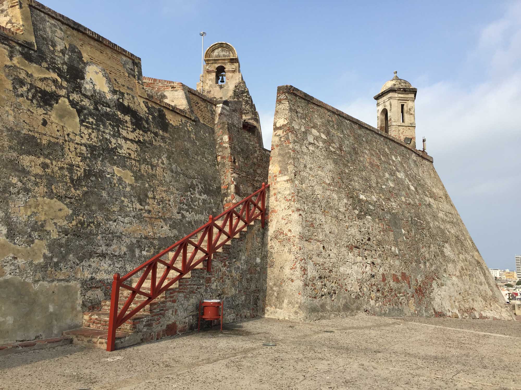 A weathered fortification with massive stone walls featuring a prominent bell tower and a bright red metal staircase leading up the exterior. The structure shows significant age and patina, typical of colonial-era coastal defense forts.