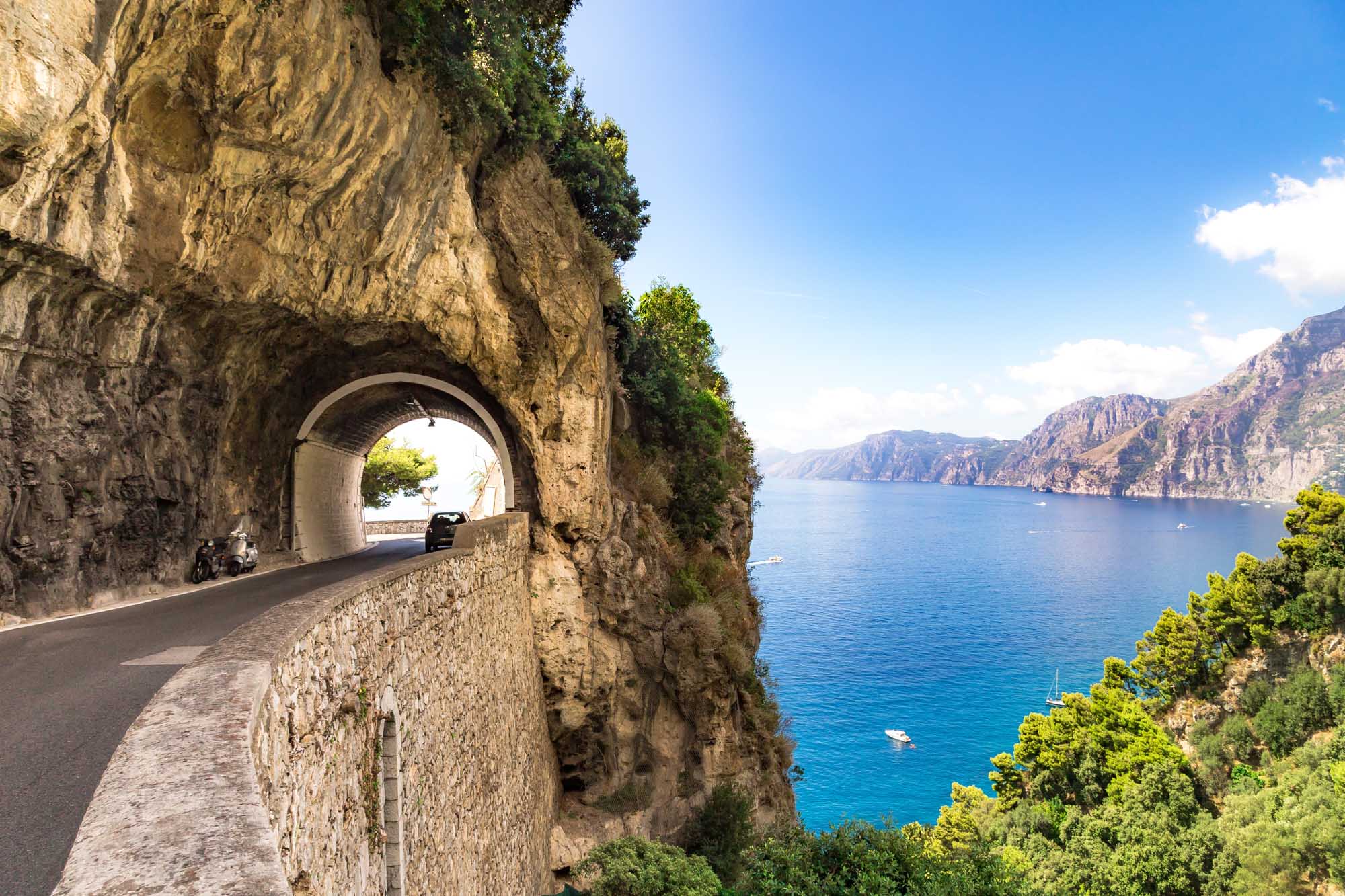 Historic Tunnel on the Amalfi Coast A scenic coastal road carved through a rocky cliff face features a stone tunnel with a scenic Mediterranean vista visible through its arch. The turquoise waters, dramatic cliff formations, and lush Mediterranean vegetation create a stunning backdrop along Italy's famous Amalfi Coast.