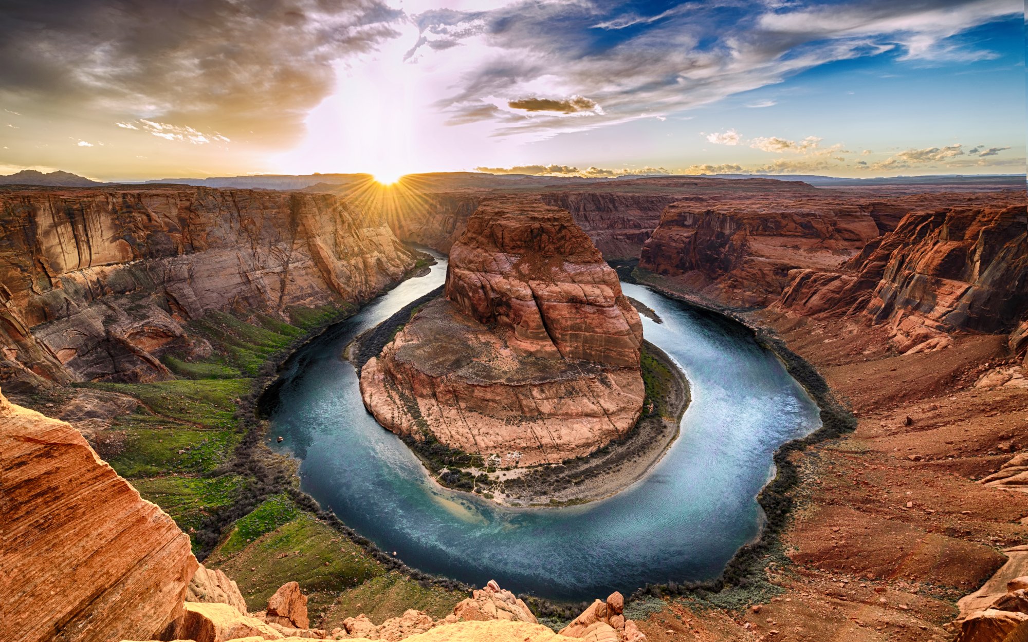 A breathtaking aerial view of Horseshoe Bend, where the Colorado River makes a dramatic 270-degree horseshoe-shaped meander through towering red sandstone cliffs. The scene captures the turquoise river snaking through the desert landscape during golden hour, with vibrant blue sky and puffy clouds creating a stunning contrast against the warm, rust-colored canyon walls.