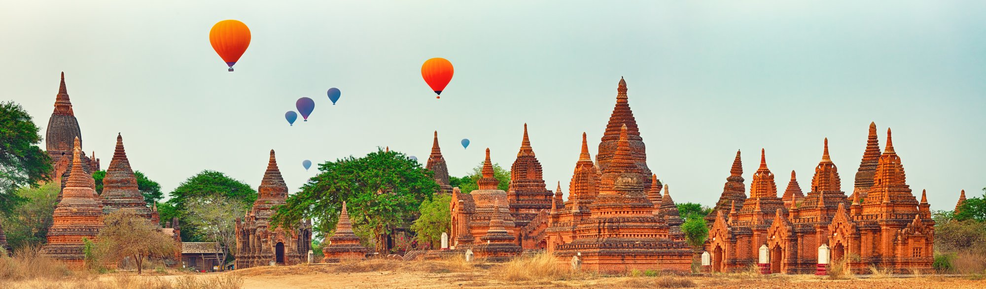 A stunning panoramic view of multiple ancient Buddhist temples with distinctive brick spires dotting a landscape, with colorful hot air balloons floating gracefully in the sky above. The scene captures the magical atmosphere of a popular Southeast Asian destination during sunrise or early morning.