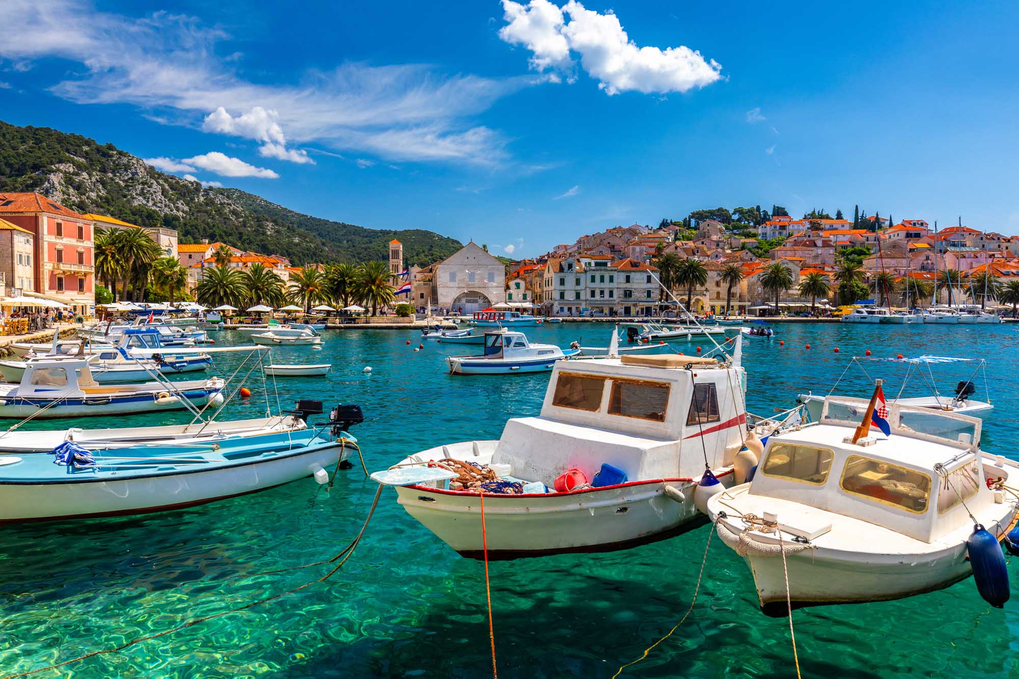 A picturesque Mediterranean harbor scene featuring numerous fishing boats anchored in crystal-clear turquoise waters off the coast of Hvar, Croatia. The historic town rises dramatically behind the harbor with traditional orange-roofed buildings nestled against a verdant hillside, showcasing the charming architecture and natural beauty of this popular Adriatic island destination.