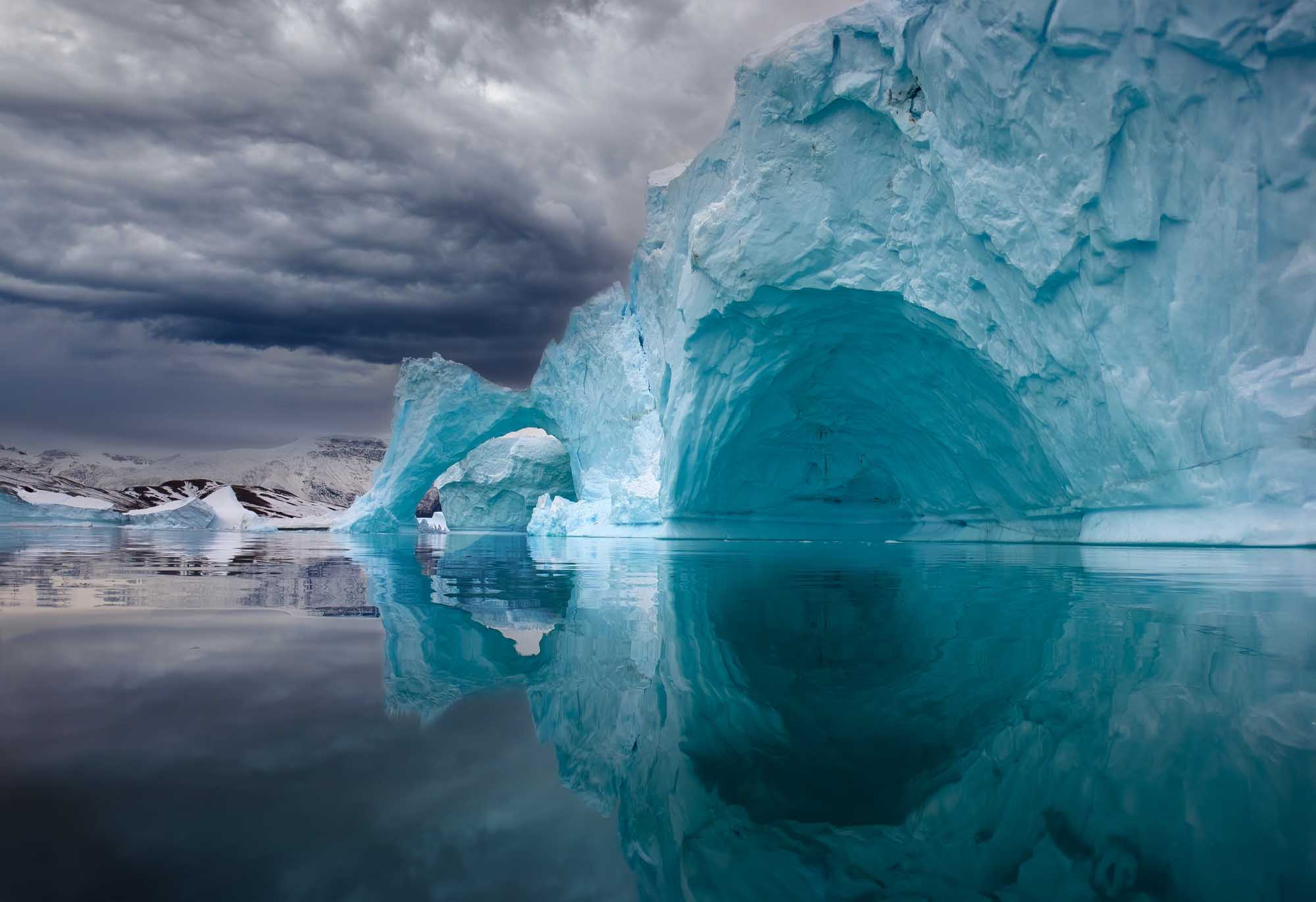 A striking turquoise iceberg featuring a natural arch formation floats in calm, mirror-like waters off the coast of East Greenland. Snow-capped mountains are visible in the background beneath a dramatic cloudy sky, creating a serene Arctic landscape.