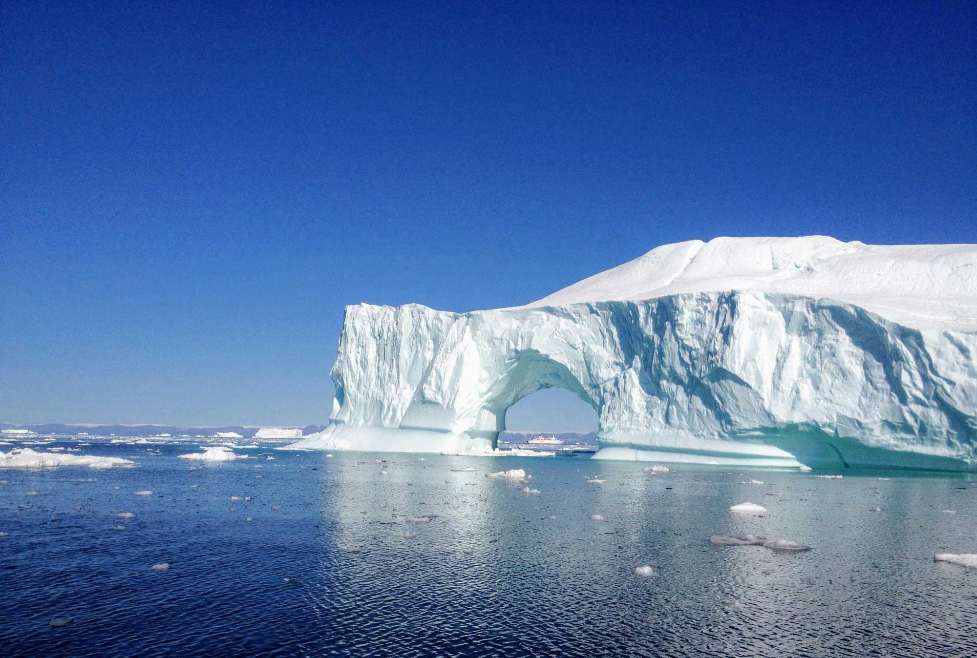 A massive tabular iceberg featuring a striking natural arch formation floats in calm Arctic waters under a clear blue sky. The ice structure displays brilliant white and turquoise hues, with smaller ice chunks scattered across the surrounding water, characteristic of polar regions during the melting season.