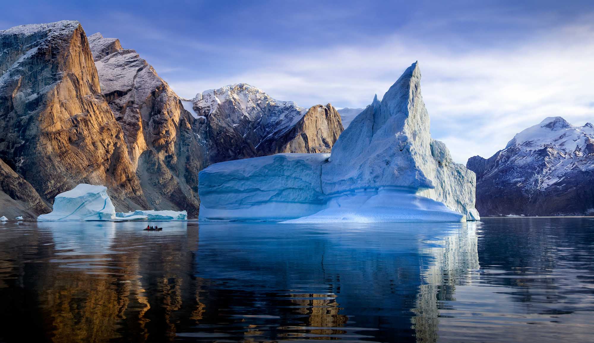 A stunning Arctic landscape featuring towering icebergs with dramatic blue and white coloring floating in calm fjord waters, surrounded by snow-capped mountains. A small boat with adventure tourists can be seen exploring among the massive ice formations in eastern Greenland.