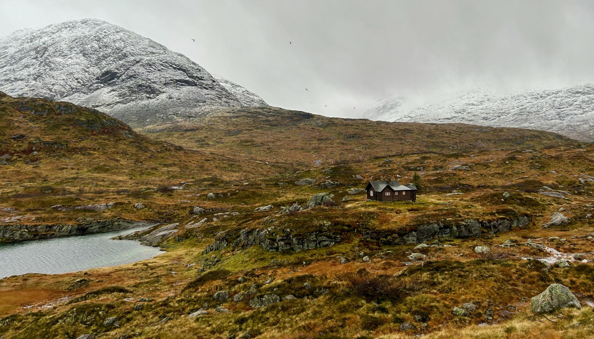 Isolated Red Cabin in Arctic Alpine Landscape A solitary red wooden cabin sits in a remote, tundra-covered valley beneath snow-capped mountains, with a small glacial pond in the foreground and sparse vegetation characteristic of Arctic or subarctic terrain.