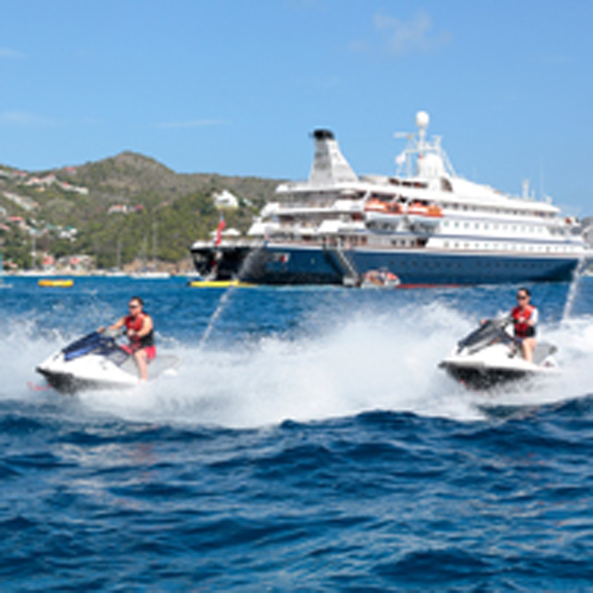 Two jet skiers in red life vests enjoy water sports in front of a large cruise ship anchored in a tropical bay, with a forested hillside visible in the background under clear blue skies.