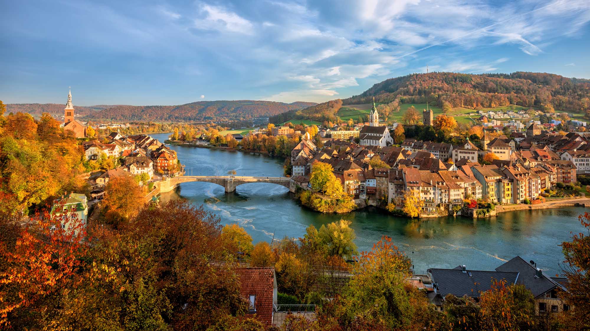 A picturesque autumn view of Laufenburg's historic old town straddling the Rhine River on the Swiss-German border, featuring traditional architecture, a stone bridge, church spires, and colorful fall foliage reflected in the blue waters below, with forested hills rising in the background.