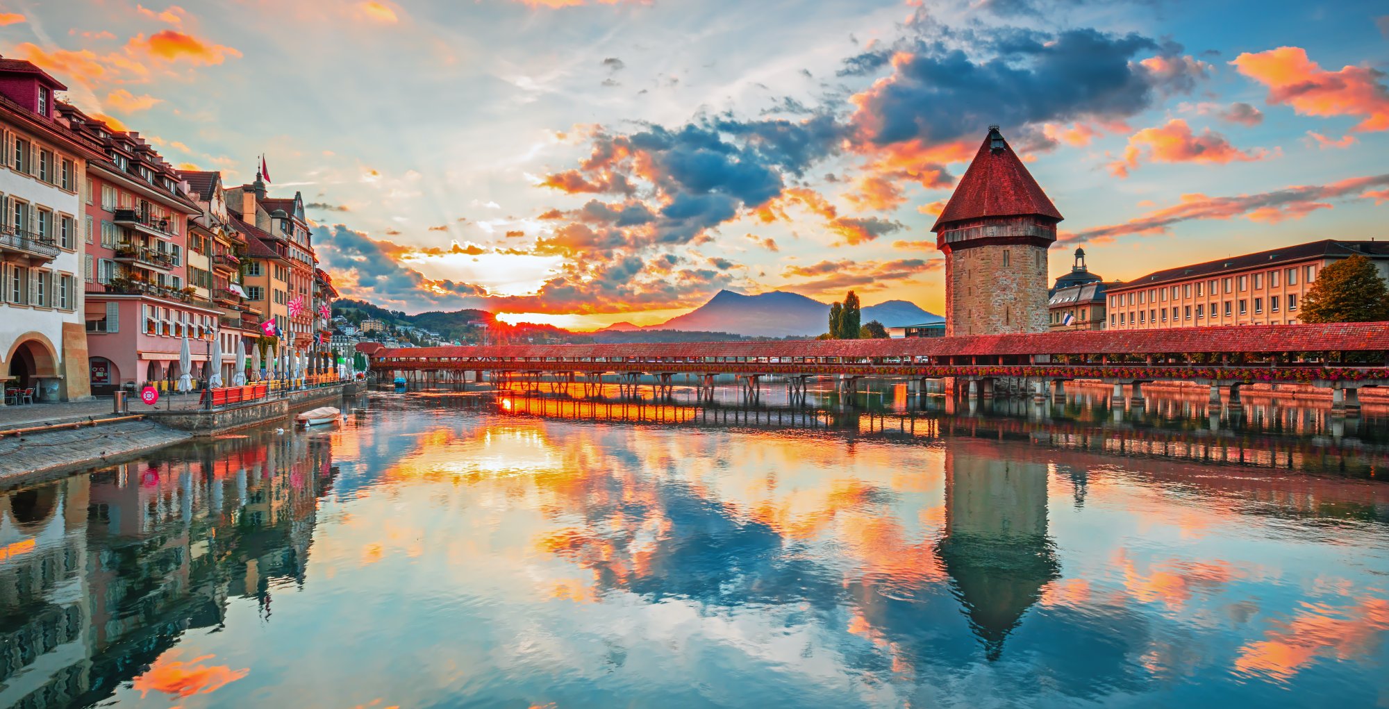 A stunning sunset view of Lucerne, Switzerland, featuring the iconic wooden Chapel Bridge (Kapellbrucke) with its distinctive red roof tower, colorful medieval buildings reflected in the calm waters of Lake Lucerne, and snow-capped mountains silhouetted against a vibrant golden and blue sky.