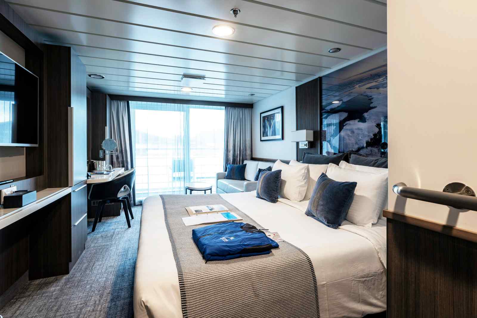 A luxurious cruise ship stateroom featuring a king-size bed with premium bedding, floor-to-ceiling windows with ocean views, modern furnishings in dark wood tones, a work desk, and sophisticated blue and white color scheme accents throughout the elegantly appointed cabin.