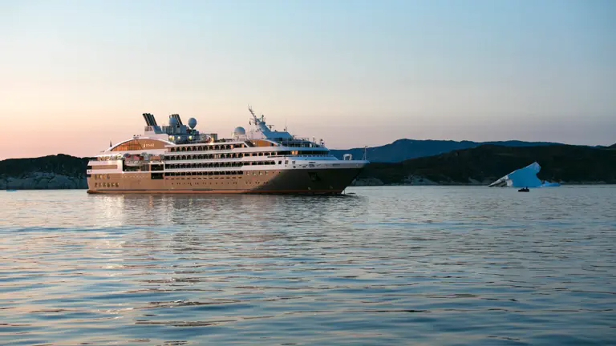 Luxury Cruise Ship in Arctic Waters A modern expedition cruise ship anchors in calm Arctic waters near Bisko Bay, with snow-capped mountains and an iceberg visible in the background during twilight hours. The vessel features a distinctive design with multiple decks and contemporary architecture typical of luxury expedition cruising.