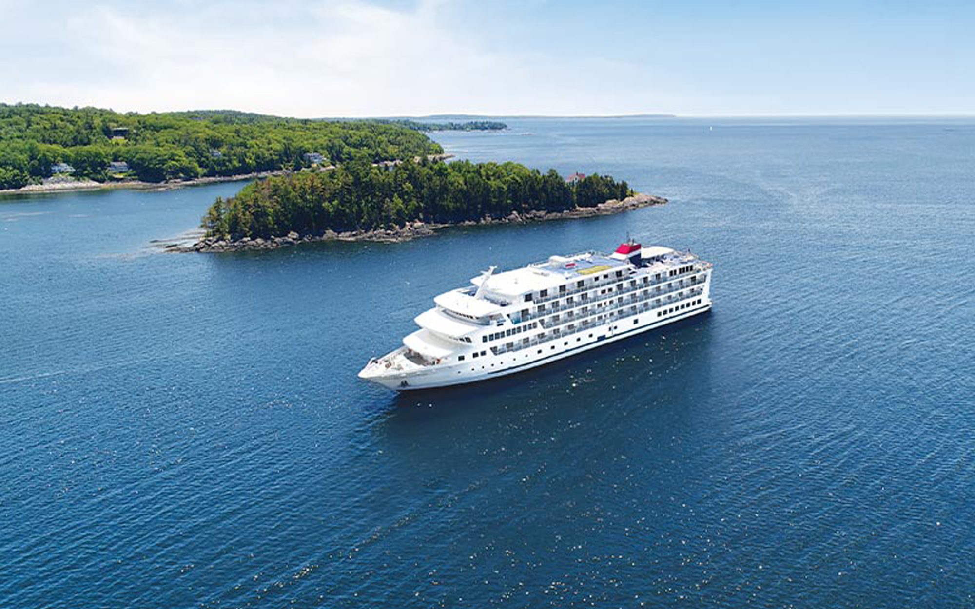 A large, modern cruise ship with distinctive white hull and red funnel anchors in the calm blue waters of Puget Sound, with a forested peninsula dotted with residential homes visible in the background. The clear day and pristine waters showcase the natural beauty of the Pacific Northwest coastal region.