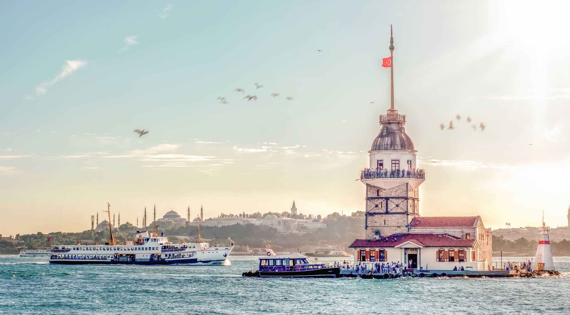 Maiden’s Tower Istanbul with Ferry Boats A scenic view of Istanbul's iconic Maiden's Tower (Kiz Kulesi) in the Bosphorus Strait, with a blue and white passenger ferry approaching the tower and the cityscape of Istanbul visible in the background under a clear sky. The historic lighthouse-style tower features a domed top with Turkish flag, surrounded by water taxis and sightseeing vessels.