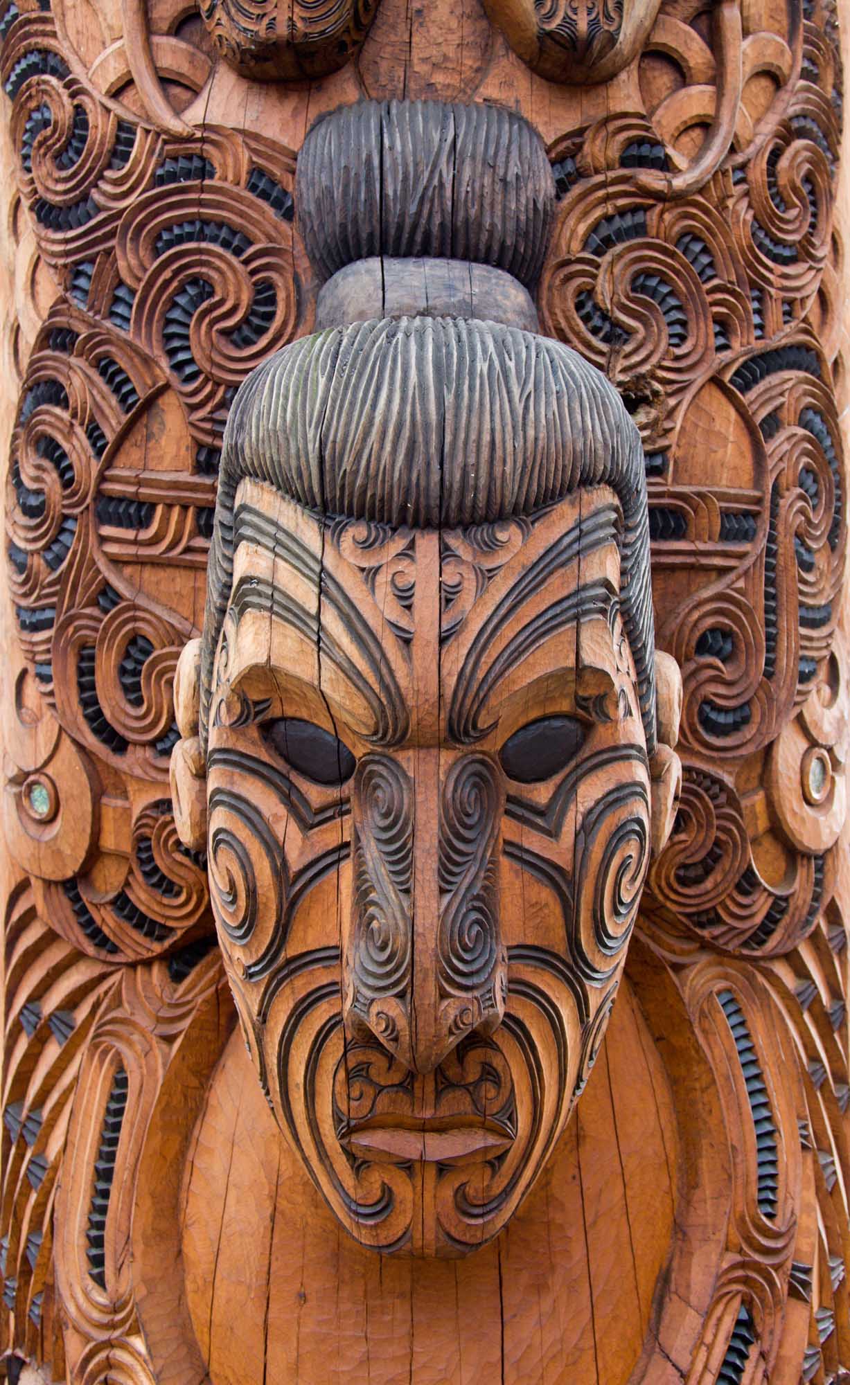An intricately carved Maori wooden face featuring traditional geometric patterns, spiral designs, and a prominent top knot hairstyle. The carving displays characteristic Maori artistic motifs with detailed facial markings and elaborate decorative elements in warm wood tones.