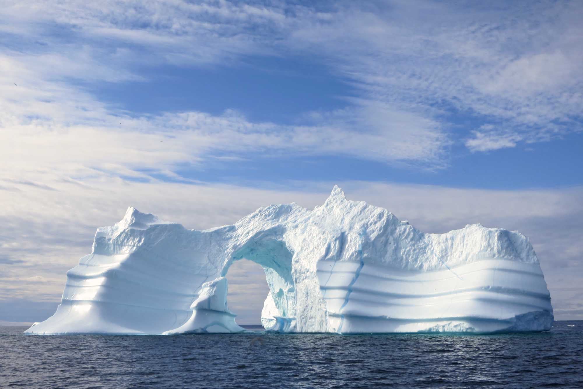 Massive Arch Iceberg in Greenland Waters A striking natural arch iceberg floats in the dark waters of Greenland, featuring brilliant white ice sculpted by wind and waves into a distinctive tunnel-like formation. The dramatic ice formation is photographed against a partly cloudy blue sky, showcasing the raw beauty of Arctic glacial landscapes.