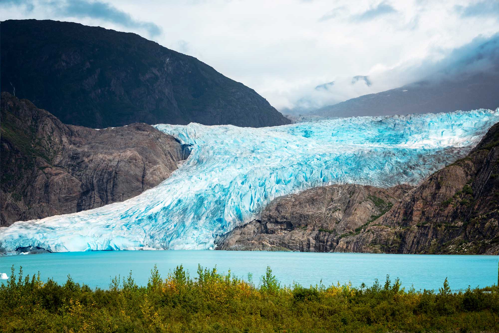 Mendenhall Glacier in Juneau, Alaska A stunning turquoise glacier cascades into a pristine glacial lake, surrounded by dramatic dark mountains and dense evergreen forest. The brilliant blue ice formations of Mendenhall Glacier showcase the raw beauty of Alaska's pristine wilderness.