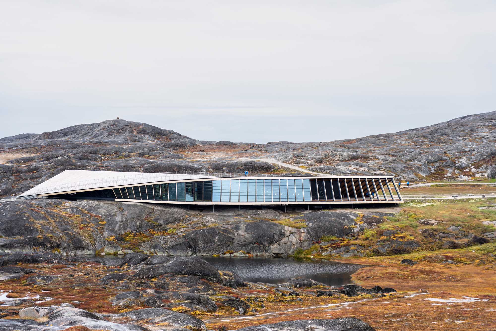 A striking contemporary building with a curved white roof and extensive blue-tinted glass panels sits atop rocky Arctic terrain in Greenland. The structure's sleek design contrasts dramatically with the barren, moss-covered landscape and distant mountains, exemplifying innovative architecture in one of the world's most remote regions.