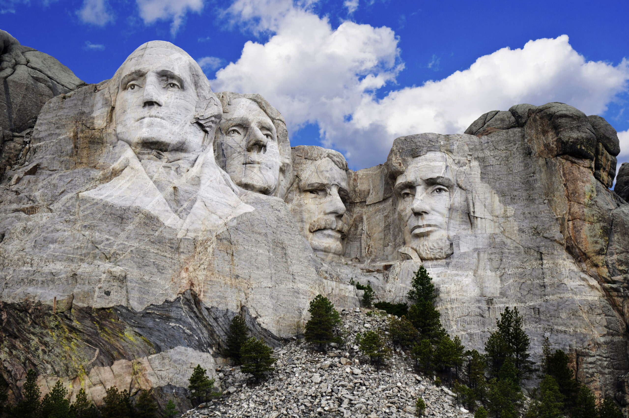 The iconic Mount Rushmore monument featuring four carved presidential faces (Washington, Jefferson, Theodore Roosevelt, and Lincoln) carved into granite cliffs against a brilliant blue sky with white clouds. Evergreen trees dot the base of the mountain.