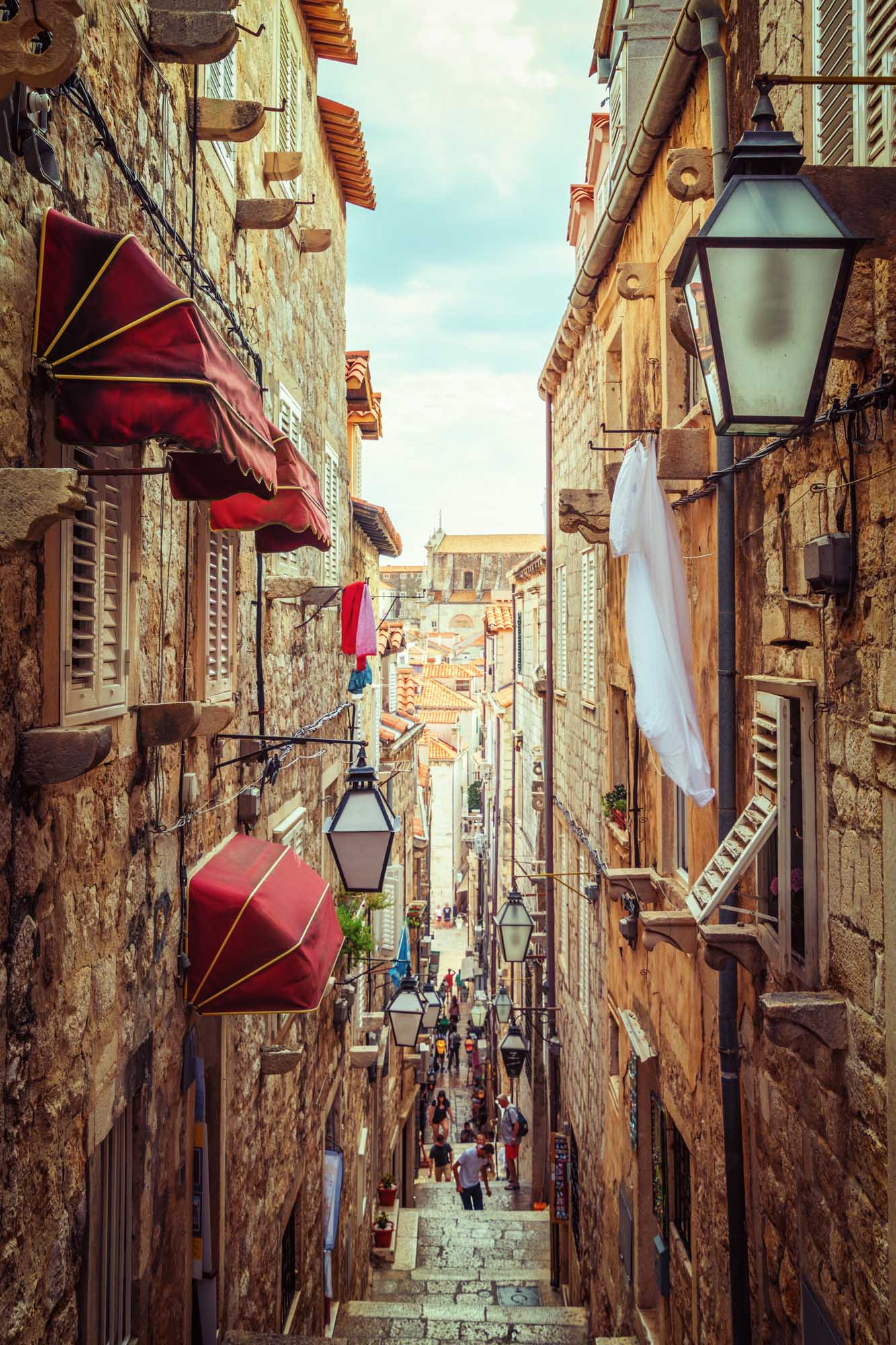 Narrow Alley in Dubrovnik Old Town A picturesque narrow stone alley in Dubrovnik's UNESCO-listed Old Town, lined with traditional architecture, red awnings, vintage lanterns, and white laundry hanging between buildings. Tourists stroll through the charming medieval street with terracotta roofs visible in the background under a clear blue sky.