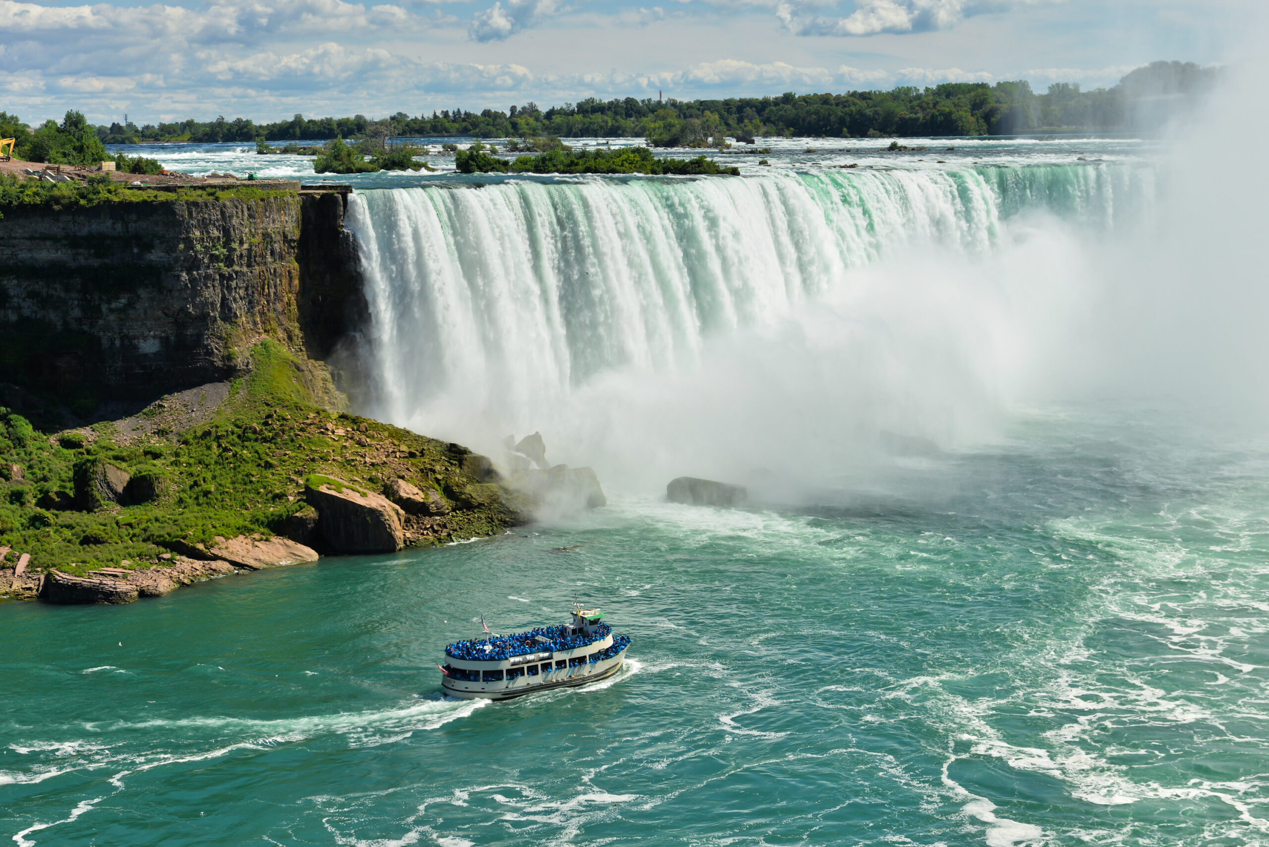 A scenic view of Niagara Falls' Horseshoe Falls with a passenger tour boat navigating the turquoise waters below, surrounded by mist and lush greenery on the Canadian side. The iconic natural wonder showcases the massive cascade of water with a blue-sky backdrop and forested landscape.
