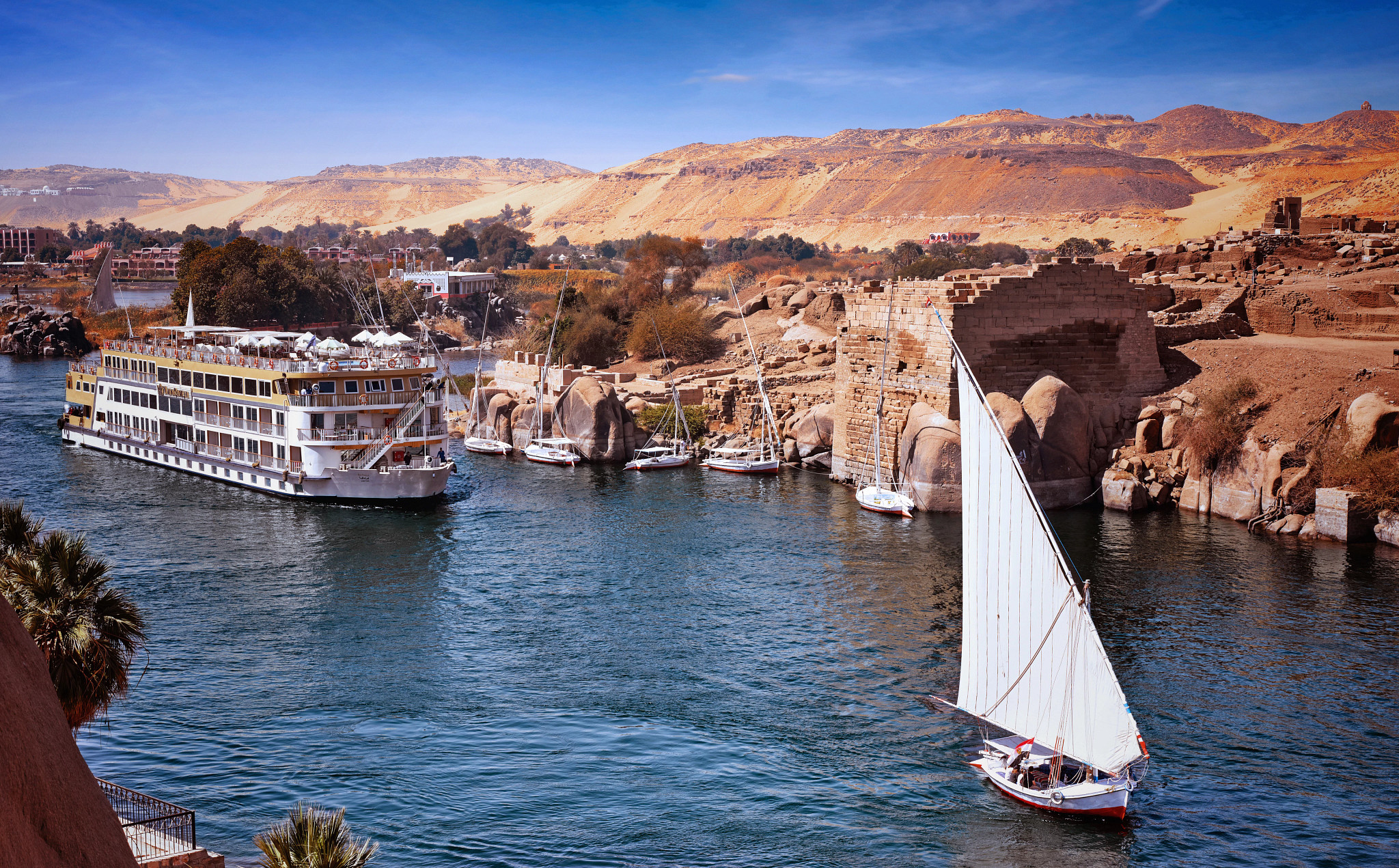 A large river cruise ship docked along the Nile River at Aswan, Egypt, with traditional felucca sailboats and ancient stone ruins visible along the riverbank.