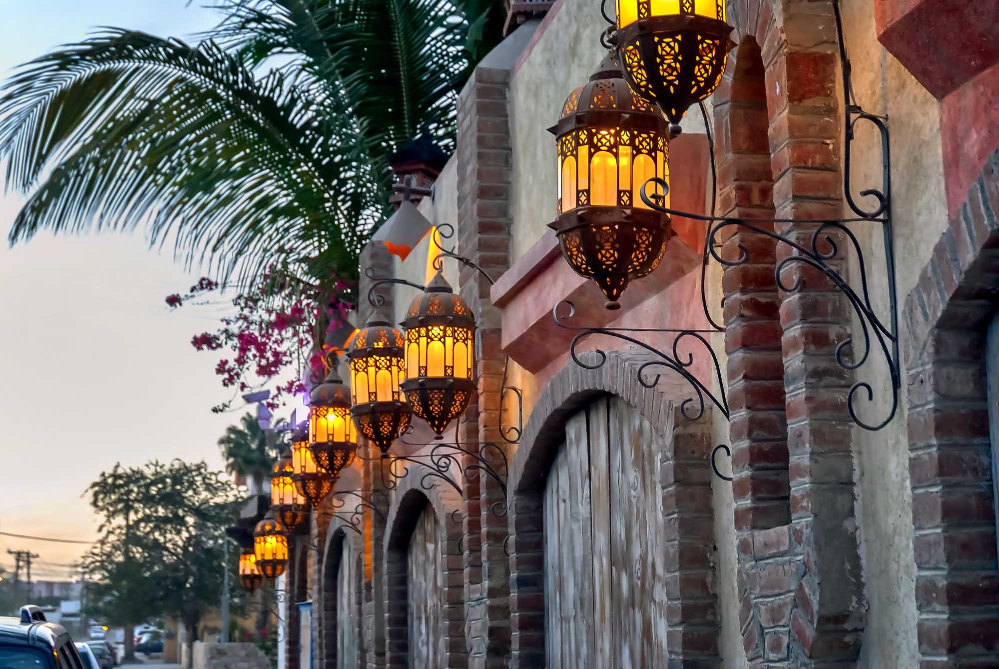 Ornate Lanterns on Historic Building in Cabo San Lucas Decorative Moroccan-style hanging lanterns with intricate metalwork and golden glass illuminate the brick facade of a historic building in Cabo San Lucas at dusk. Vibrant bougainvillea flowers and a palm tree frame the architectural details, creating a warm, inviting ambiance typical of this popular coastal destination.
