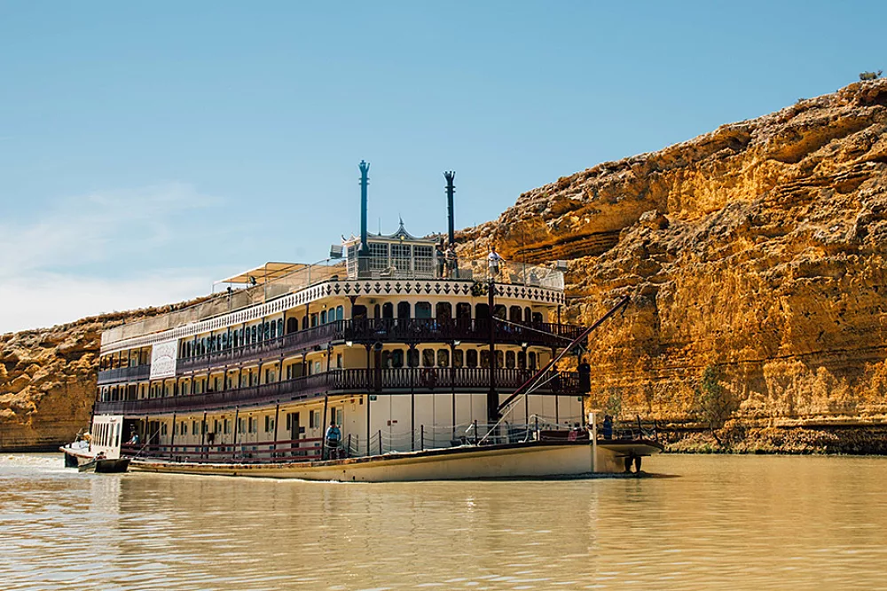 A classic multi-deck paddlewheel riverboat with distinctive twin smokestacks navigates a muddy river, with dramatic golden-hued limestone cliffs towering in the background. The vessel features traditional stern-wheel design with ornate white railings and multiple passenger decks typical of Mississippi River-style cruising.