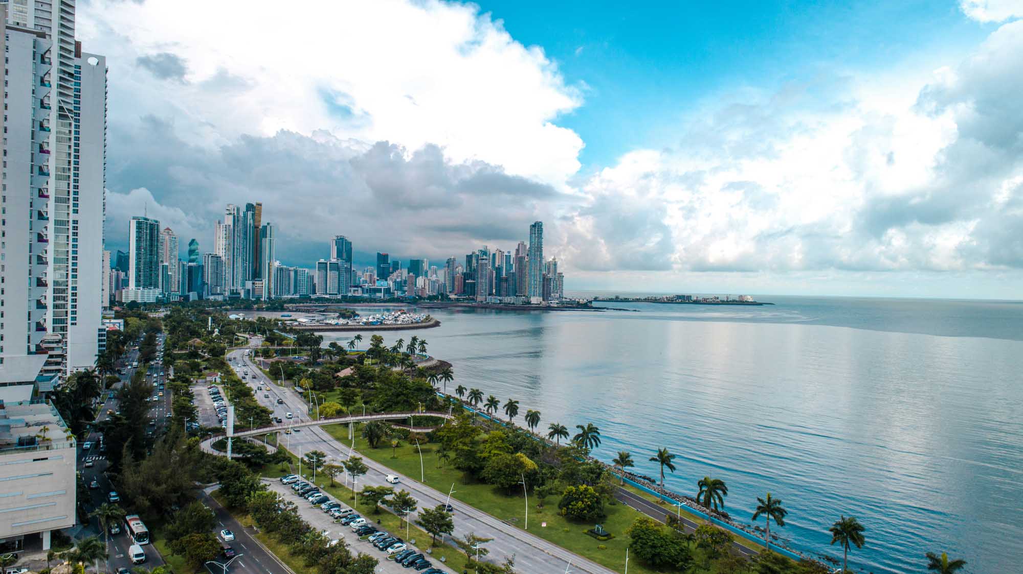 A scenic aerial view of Panama City's modern high-rise skyline along the Pacific coast, featuring contemporary residential and commercial towers, a manicured waterfront park with palm trees, and calm turquoise waters under a partly cloudy sky.
