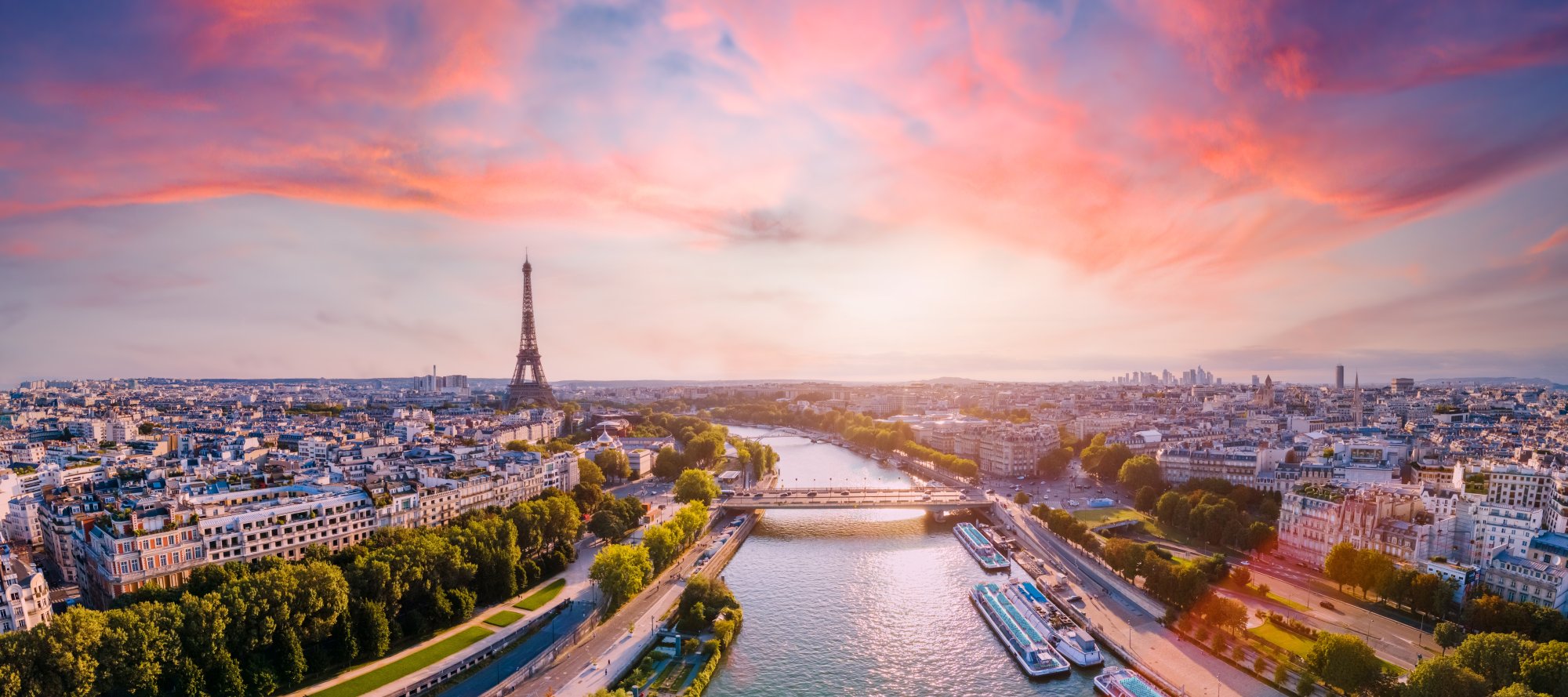 An aerial view of Paris at golden hour, featuring the iconic Eiffel Tower prominently in the center, with the Seine River winding through the cityscape, historic Haussmann-style buildings, and vibrant pink and orange sky reflecting off the water. This breathtaking sunset panorama captures the romantic essence of the French capital.