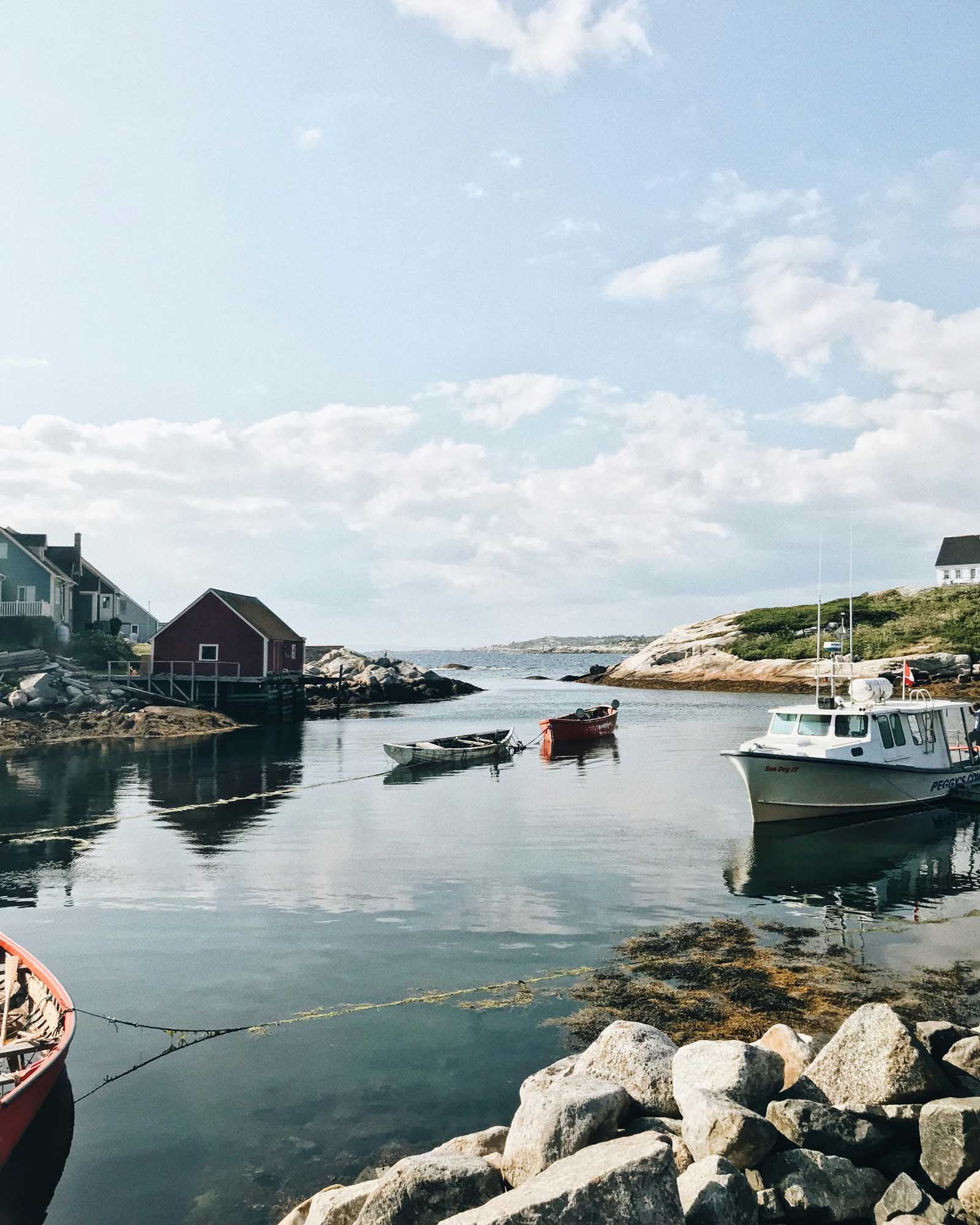 Peaceful Fishing Village Harbor with Boats A serene coastal fishing village with traditional wooden buildings, moored fishing boats, and rocky shoreline under a partly cloudy sky. The calm harbor waters reflect the picturesque New England or Maritime charm of this quiet seaside settlement.