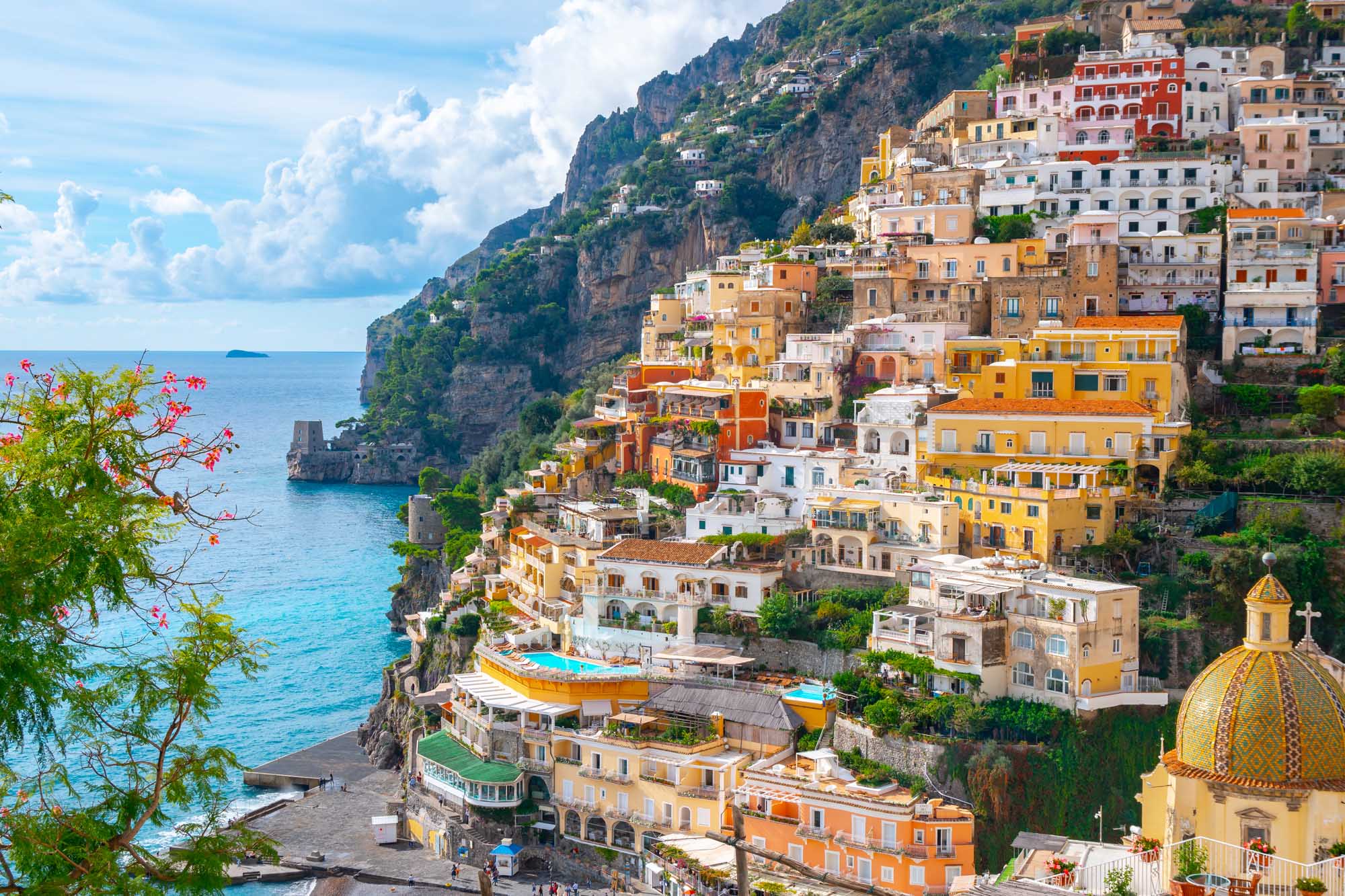 Positano Village on the Amalfi Coast A stunning coastal vista of Positano, Italy, showcasing colorful pastel-hued buildings cascading down steep cliffs to the crystalline Mediterranean Sea. The iconic yellow-domed church (Chiesa di Santa Maria Assunta) is visible on the right, with vibrant yellow, orange, pink, and white buildings stacked vertically along the dramatic rocky coastline, epitomizing the picturesque charm of the Amalfi Coast.