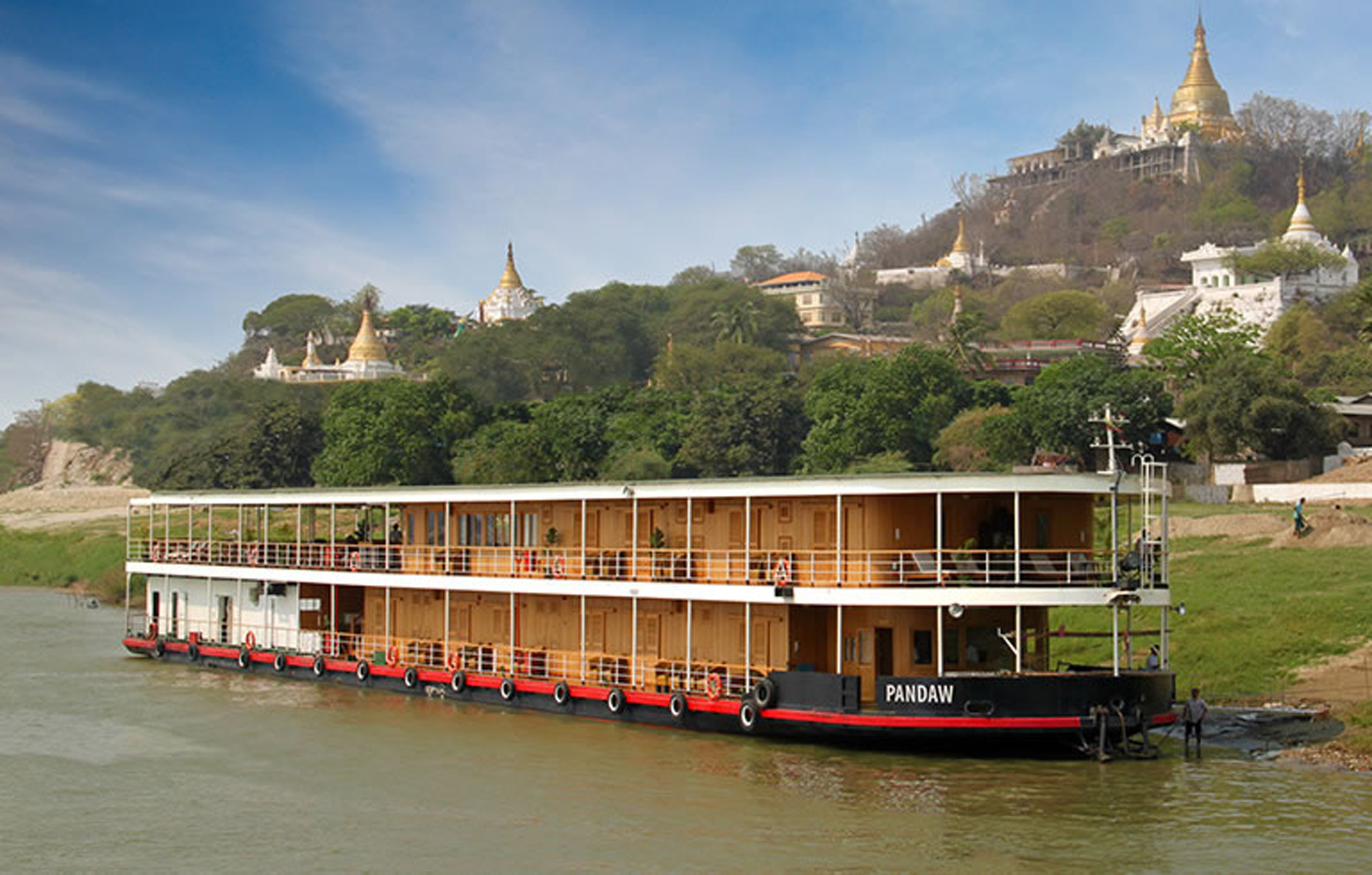 RV Pandaw River Cruise Ship on Irrawaddy River A two-deck river cruise vessel named Pandaw moored on the Irrawaddy River in Myanmar, with golden pagodas and Buddhist temples visible on the hillside in the background under clear blue skies.