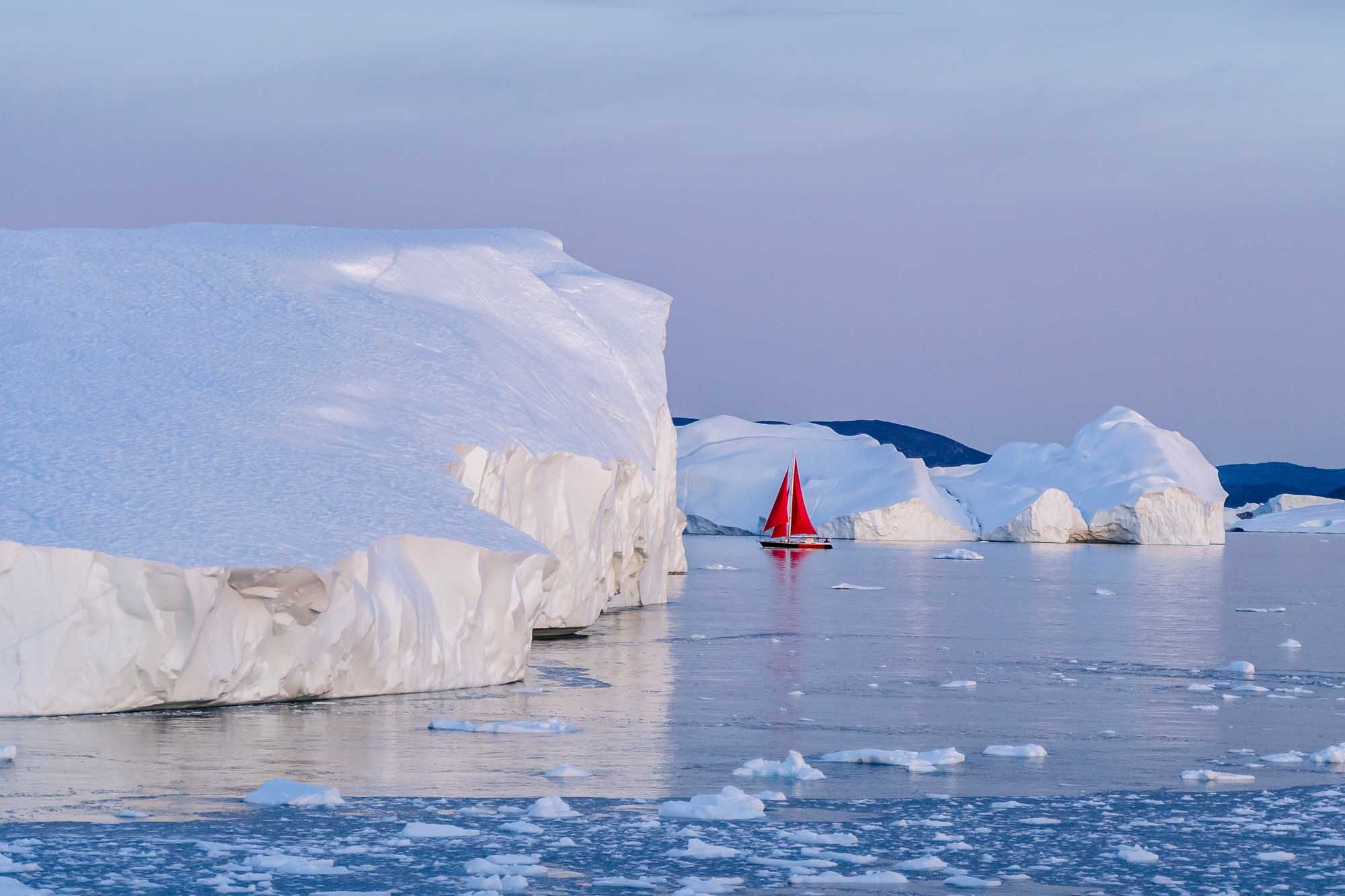 A striking red-sailed vessel navigates through a pristine Arctic landscape dotted with massive white icebergs. The scene captures the contrast between the vivid crimson sail and the pale blue waters and towering ice formations, showcasing exploration in one of Earth's most remote and dramatic polar regions.