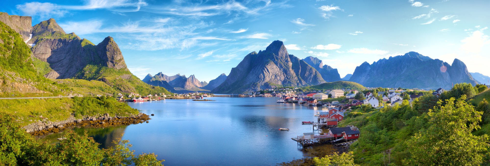A stunning panoramic view of Reine, a picturesque fishing village nestled in the Lofoten Islands of northern Norway, featuring traditional red cabins built over the water, dramatic jagged mountain peaks, and calm blue fjord waters under a clear sky.