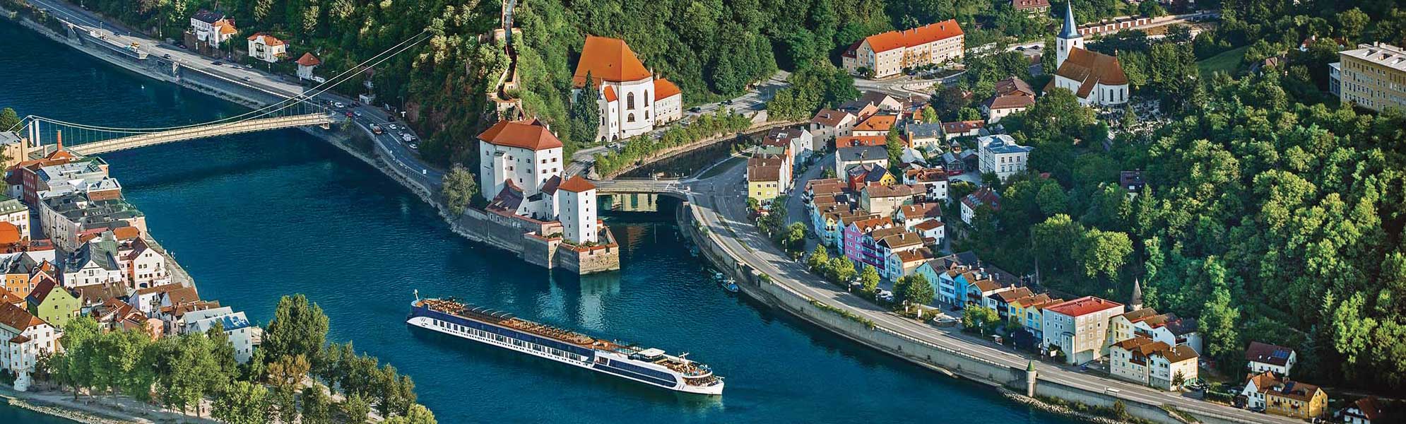 An aerial view of a river cruise ship navigating through the picturesque town of Passau, Germany, situated at the confluence of three rivers. The scene showcases the town's distinctive architecture with colorful buildings, baroque churches with orange-tiled roofs, and surrounding forested hillsides, with multiple bridges spanning the blue waters.