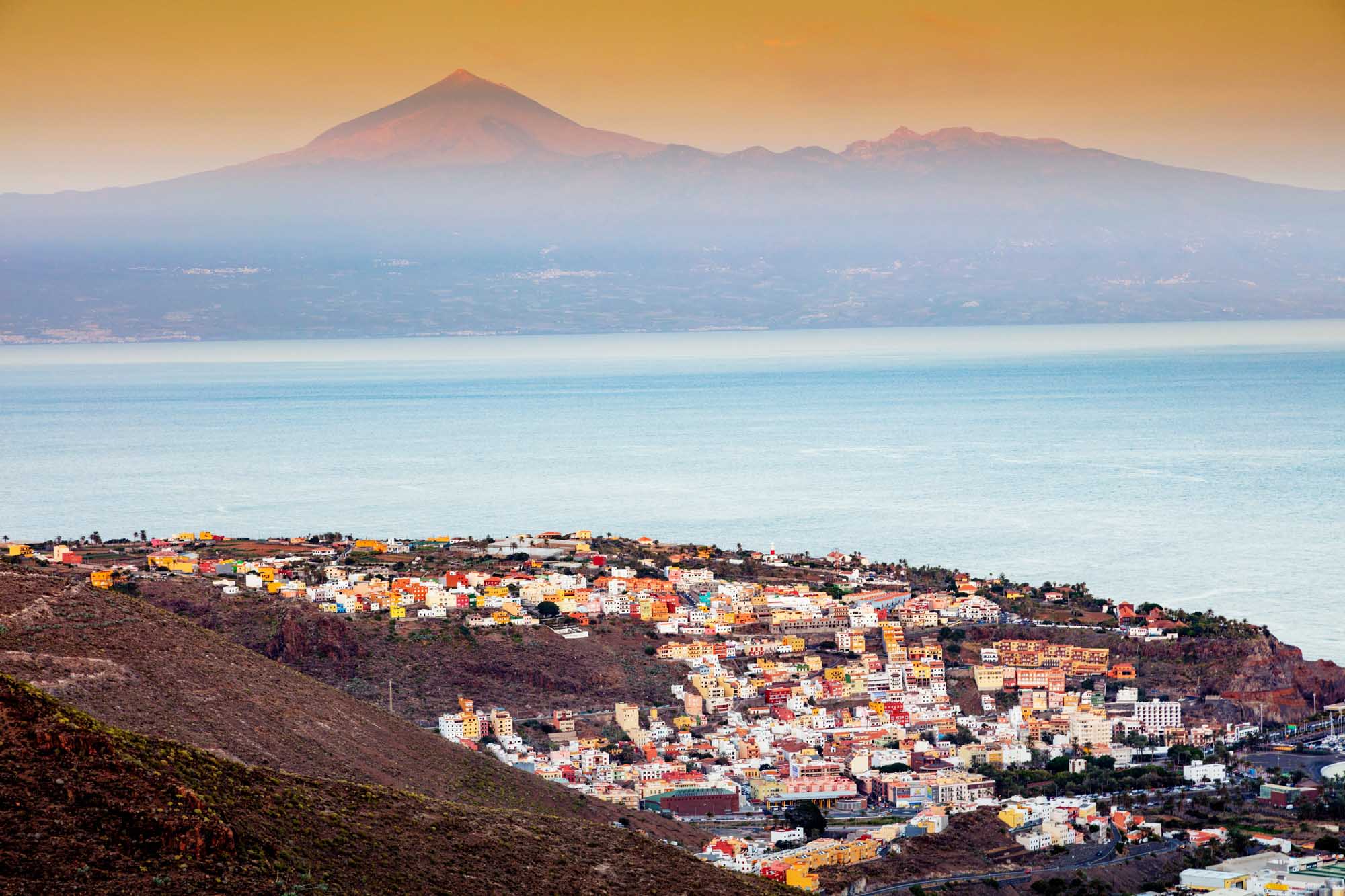 A picturesque coastal town cascades down volcanic slopes toward turquoise Atlantic waters, with its distinctive colorful buildings in vibrant reds, yellows, and whites. In the distance across the ocean, the majestic snow-capped peak of Mount Teide on Tenerife dominates the horizon under a golden sunrise sky.