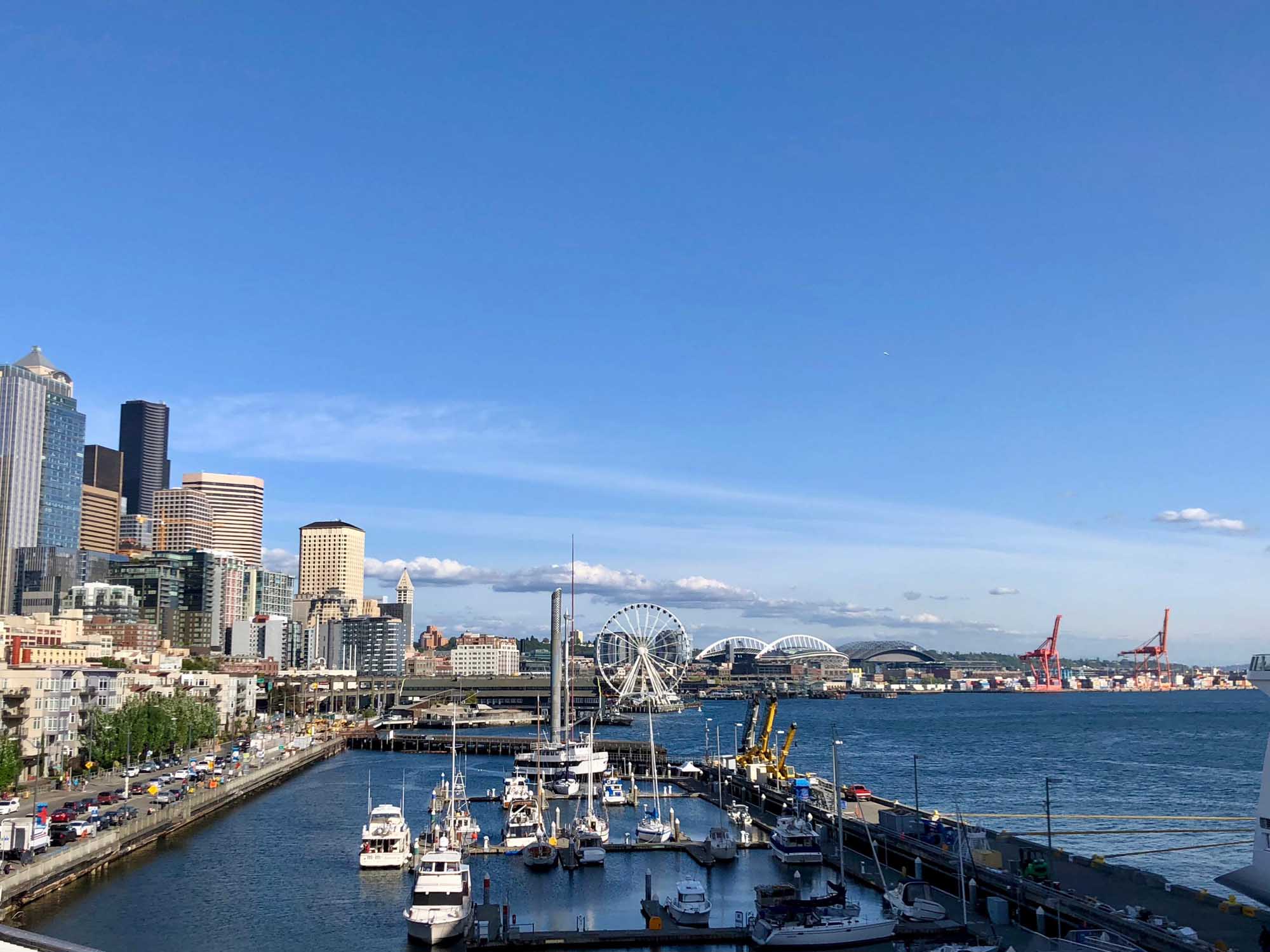 A panoramic view of Seattle's downtown skyline and waterfront featuring the iconic Space Needle, the Seattle Great Wheel, and numerous moored vessels in Elliott Bay. The image captures the bustling port district with modern skyscrapers, historic buildings, and the pristine blue waters of Puget Sound under clear skies.