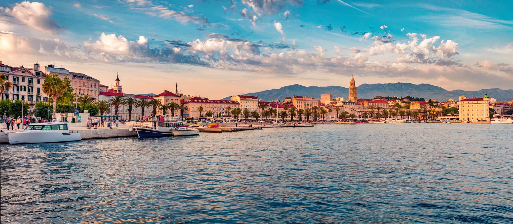 A stunning panoramic view of Split, Croatia's historic waterfront at sunset, featuring traditional Mediterranean architecture, palm-lined promenade, moored boats, and mountains in the distance under a dramatic blue sky with white clouds.