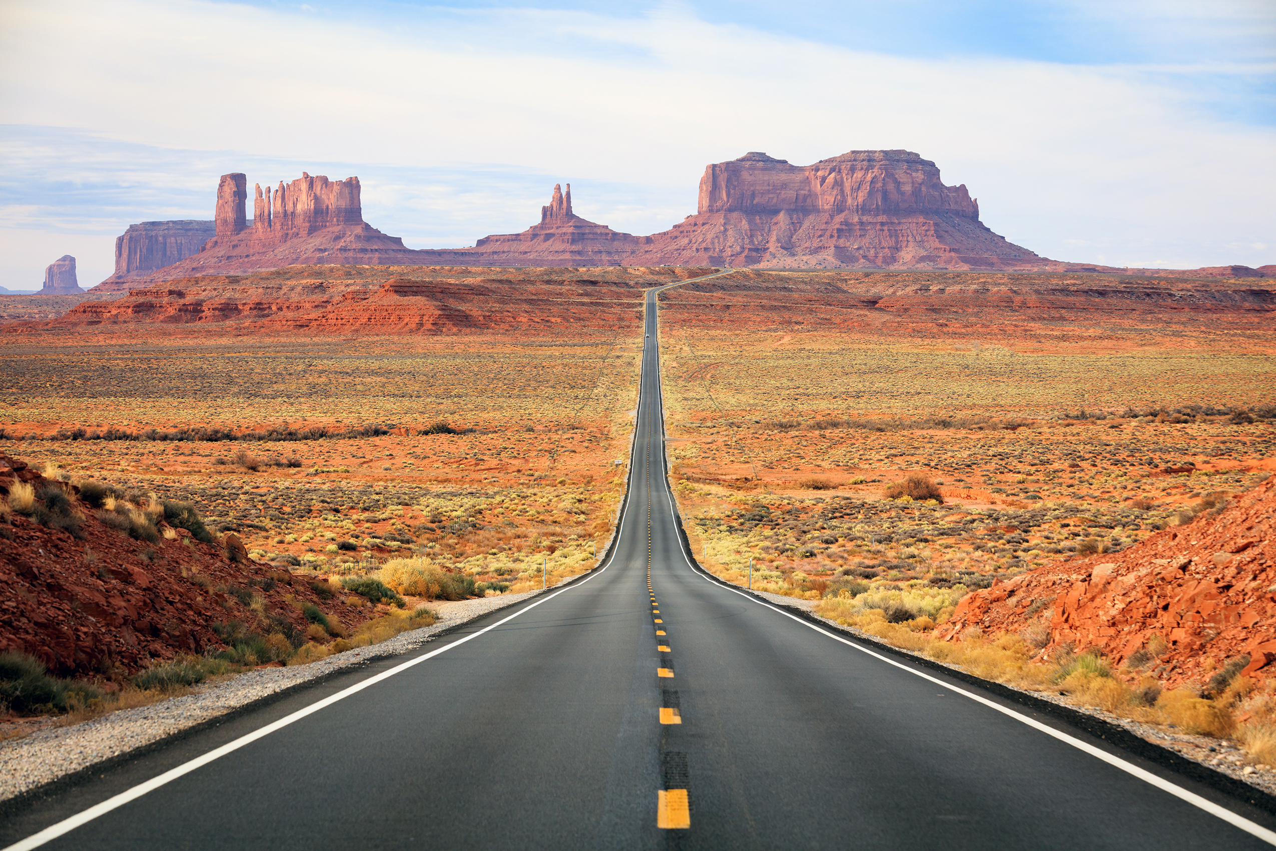 A perfectly straight asphalt highway stretches toward the horizon, leading directly to the iconic buttes and mesas of Monument Valley. The image captures the classic American Southwest landscape with striking red rock formations, desert scrubland, and a clear blue sky, exemplifying the scenic road trip experience.