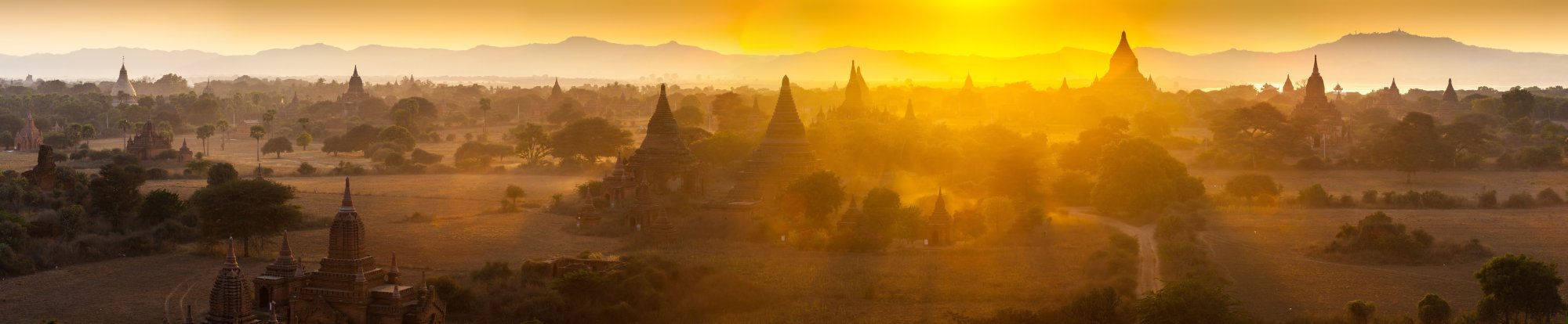 A breathtaking panoramic view of thousands of ancient Buddhist temples silhouetted against a golden sunset in Bagan, Myanmar. The landscape is bathed in warm amber light with misty atmospheric haze obscuring the lower valleys, creating a mystical and timeless scene of this UNESCO World Heritage Site.