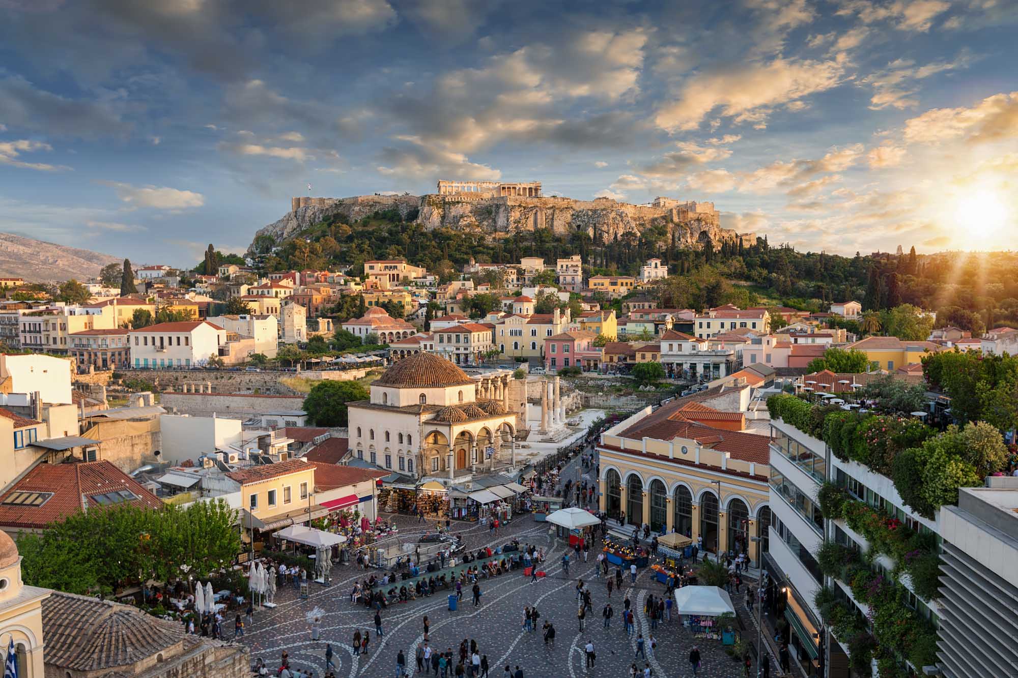 Athens Acropolis and Plaka at Golden Hour A stunning golden-hour view of Athens' historic Plaka district with colorful neoclassical buildings clustered below the illuminated Acropolis and Parthenon Temple perched atop a rocky hill. The scene captures the Byzantine Church of the Holy Apostles in the foreground with tourists gathered in the bustling square, exemplifying the blend of ancient history and vibrant modern life in Greece's capital.