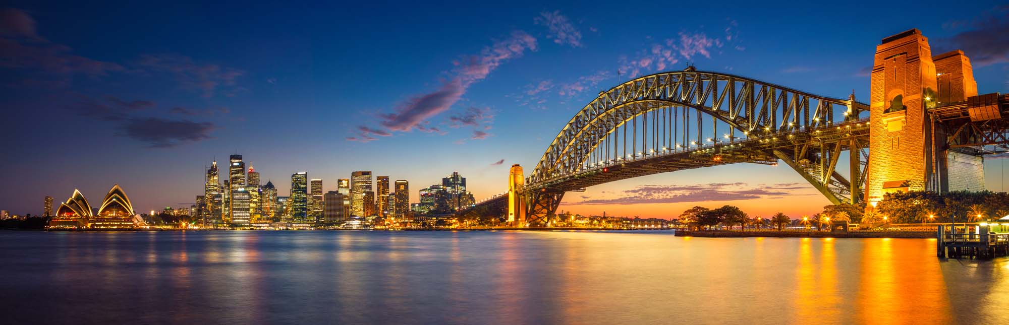 Sailing Ship in Milford Sound Fjord A stunning panoramic view of Sydney, Australia during the blue hour twilight, featuring the iconic Sydney Harbour Bridge illuminated in golden light, the distinctive Sydney Opera House on the left, and the glittering skyline reflected in the calm harbour waters.