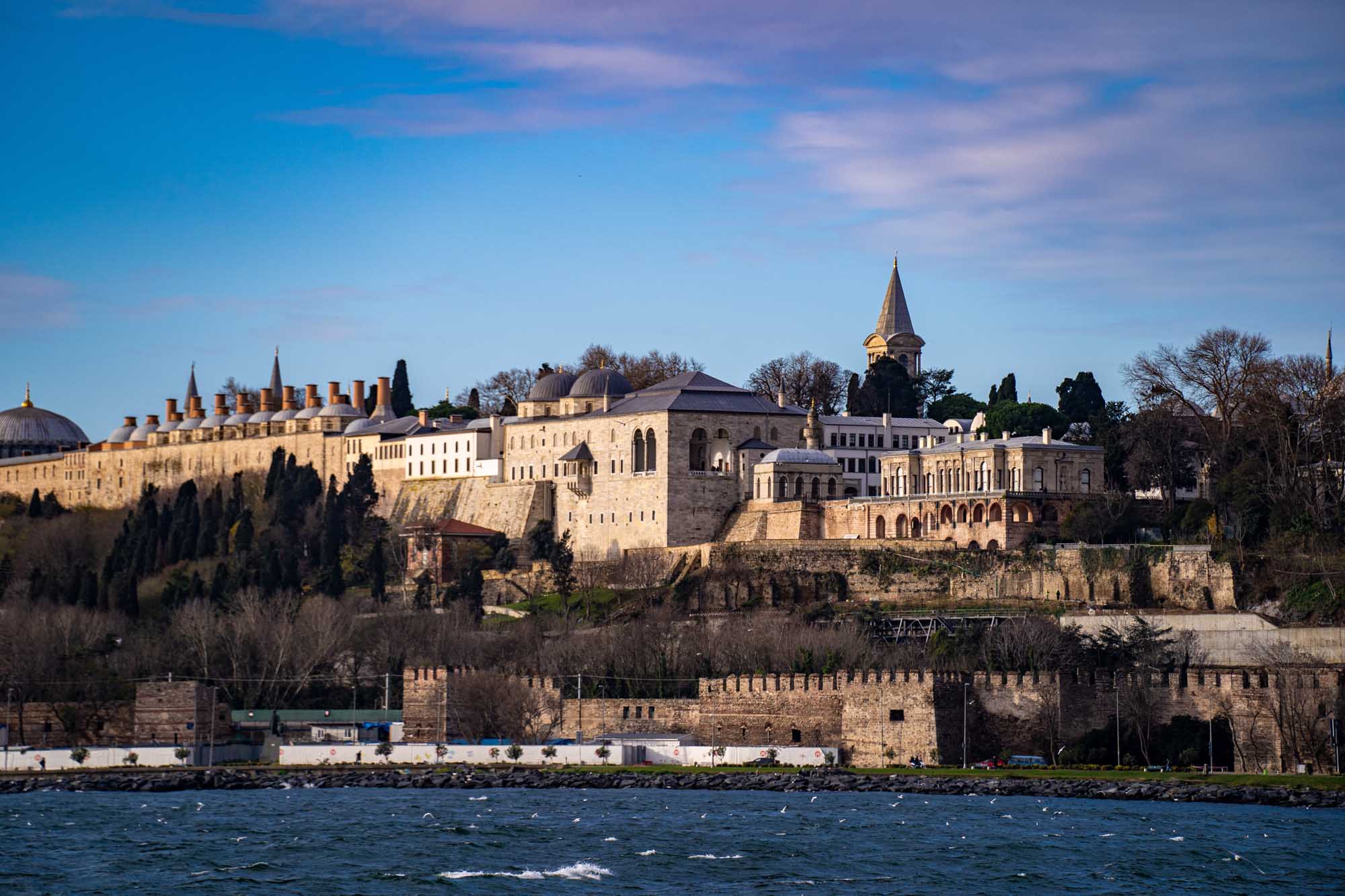 A stunning waterfront view of the historic Topkapi Palace in Istanbul, Turkey, photographed from the Bosphorus Strait. The ornate Ottoman architecture features distinctive domes, minarets, and stone fortifications set against a clear blue sky, with cypress trees framing the palatial complex on the peninsula.