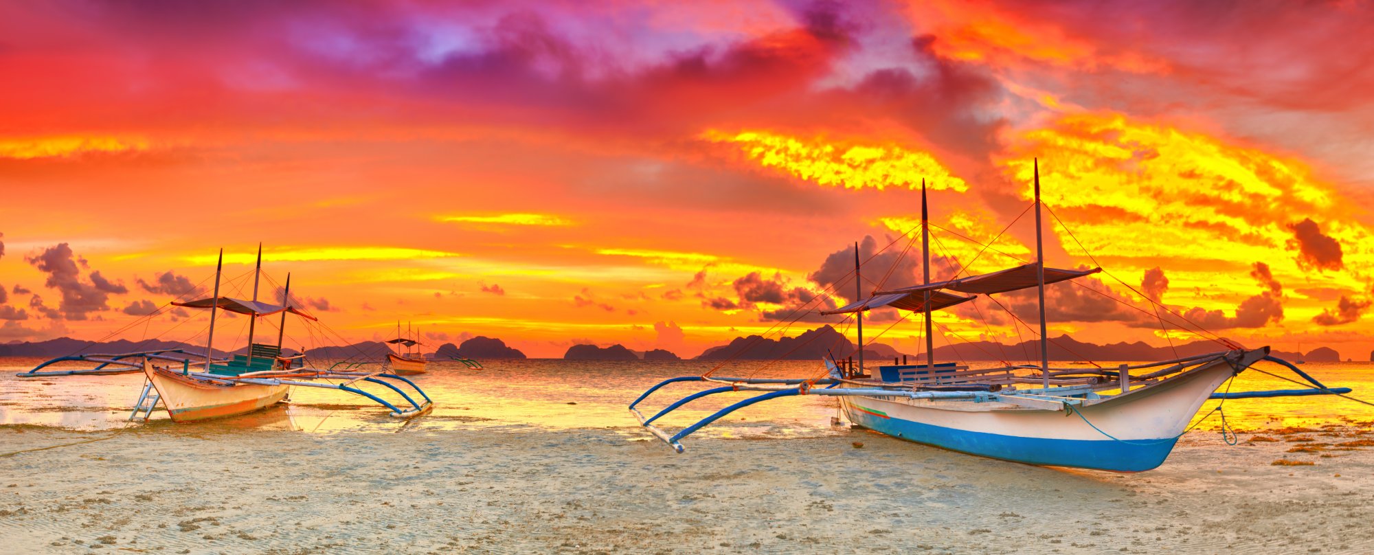 A picturesque tropical beach scene at golden hour featuring traditional Filipino outrigger boats (bancas) with distinctive blue and yellow hulls anchored in shallow waters. The dramatic sunset sky displays vibrant orange, yellow, and purple hues reflected on the calm sea, with distant limestone karsts silhouetted on the horizon, characteristic of Southeast Asian island destinations.