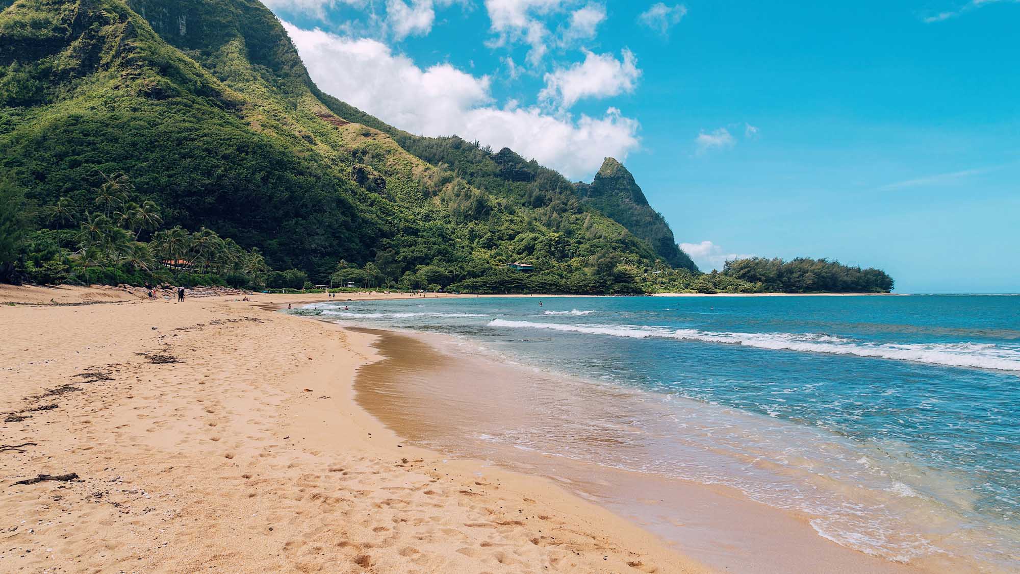 Tropical Beach Cove with Lush Mountain Backdrop A pristine sandy beach with turquoise waters and gentle waves, framed by dramatic lush green cliffs and mountains. The idyllic tropical setting features a small number of visitors enjoying the shoreline beneath a bright blue sky with scattered clouds.