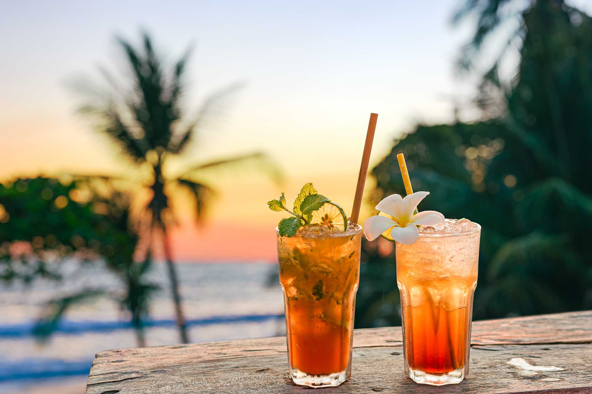 Tropical Cocktails at Sunset Beach Two vibrant tropical cocktails with orange and amber hues sit on a weathered wooden table overlooking a beach at sunset. The drinks are garnished with fresh mint, lime slices, and white flowers, with palm trees and ocean waves visible in the soft-focus background during golden hour.